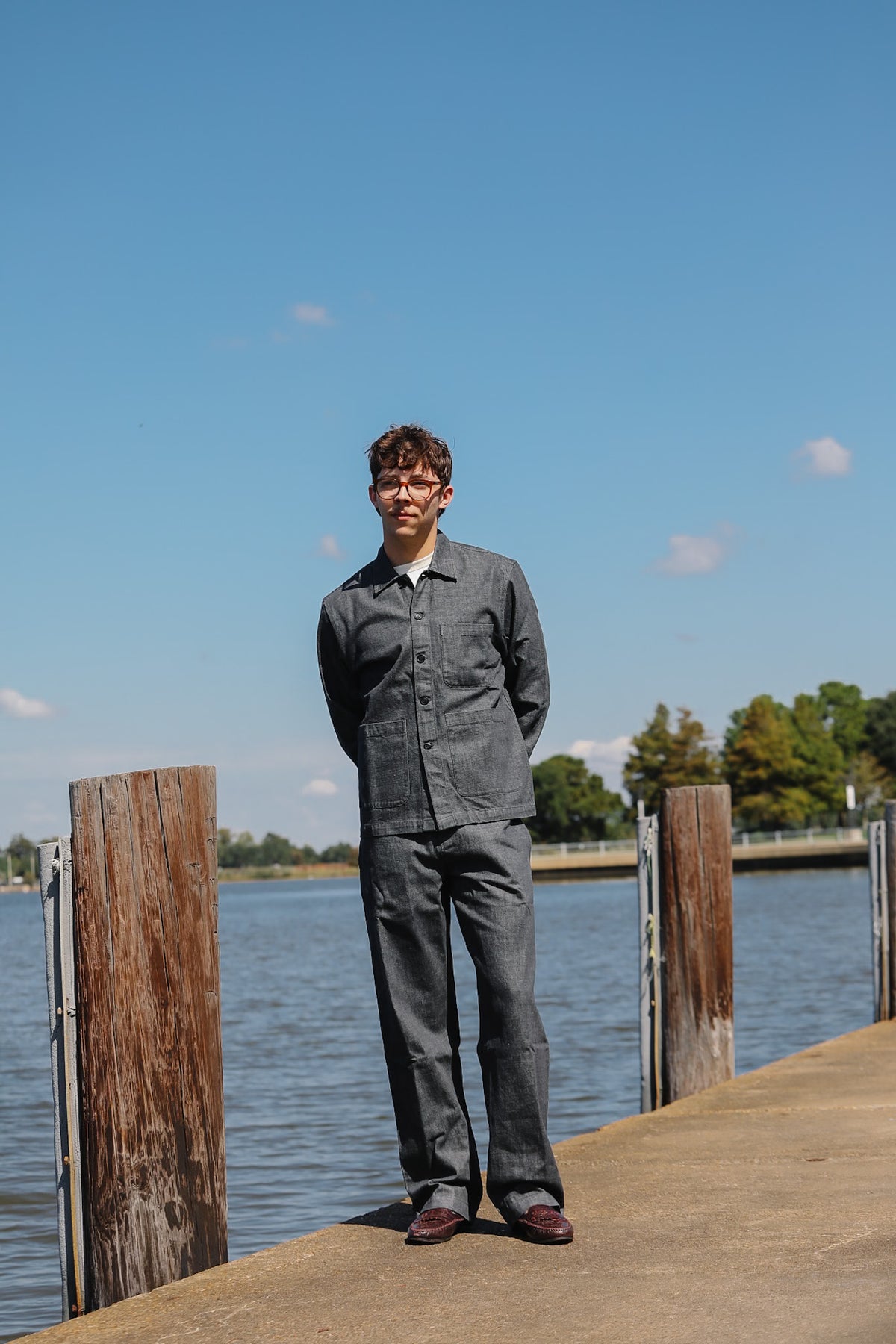 A man wearing a matching gray two-piece work uniform stands on a concrete dock next to a body of water under a clear blue sky.