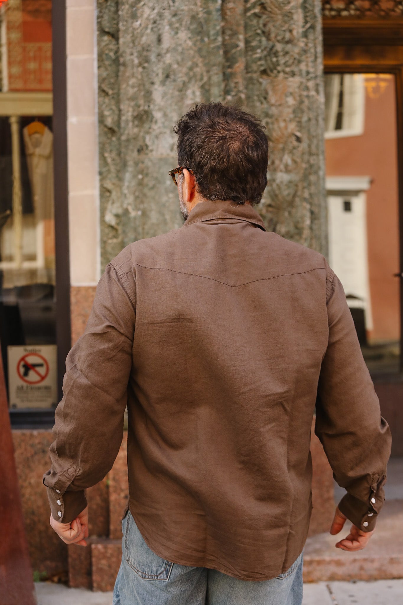 Man wearing a brown leather jacket walking away from the camera with a tree and building in the background.