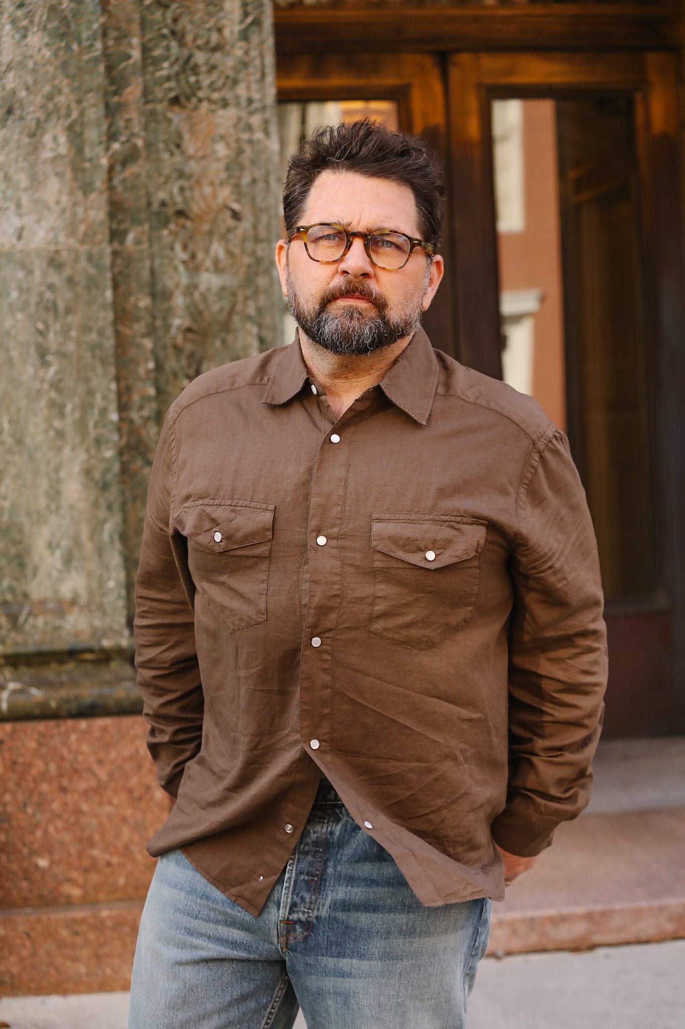 Man wearing a brown shirt and glasses standing against a building.
