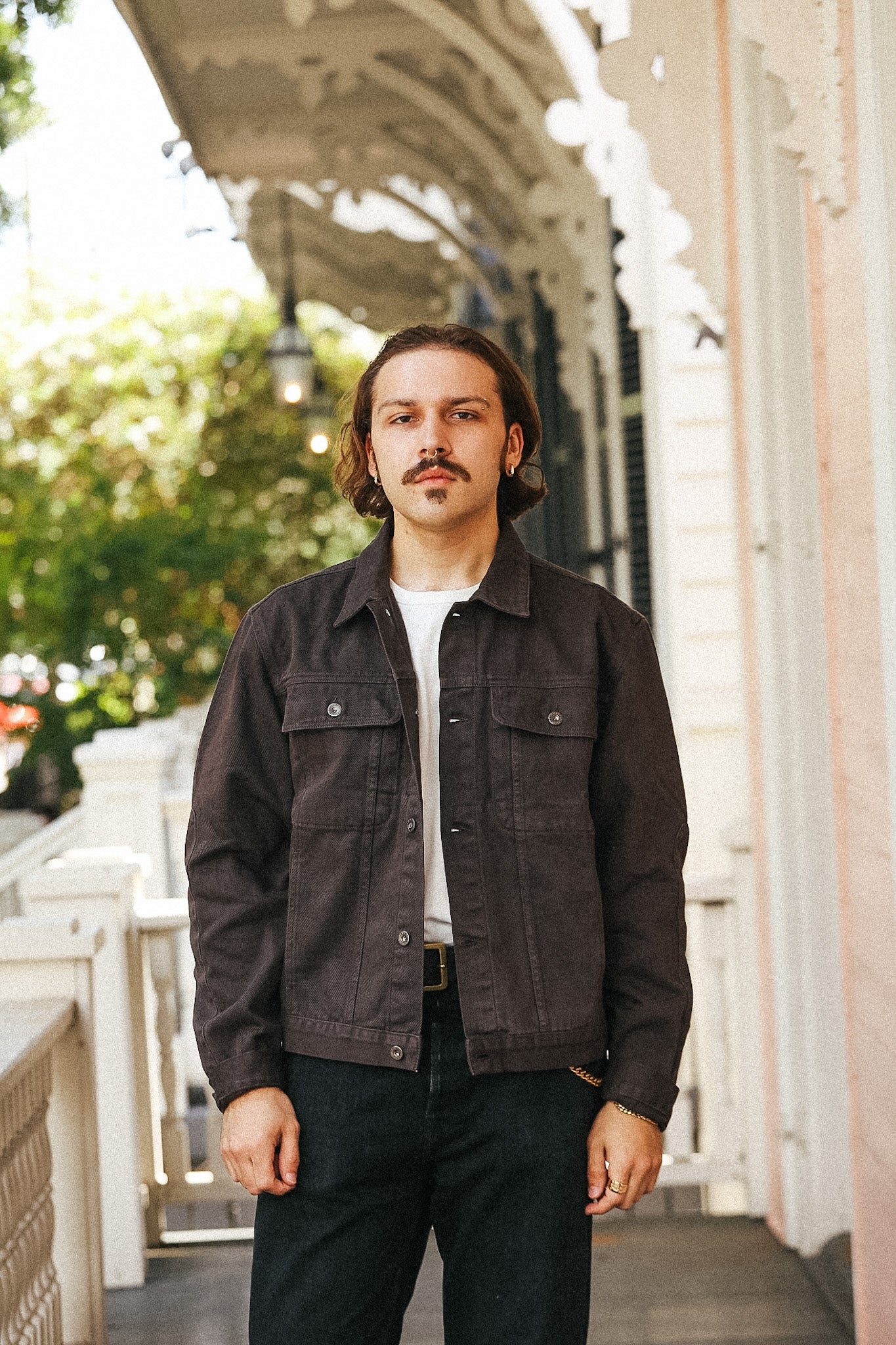 A man with a mustache stands on a porch wearing a dark brown utility jacket over a white t-shirt and dark jeans.