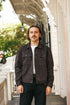 A man with a mustache stands on a porch wearing a dark brown utility jacket over a white t-shirt and dark jeans.