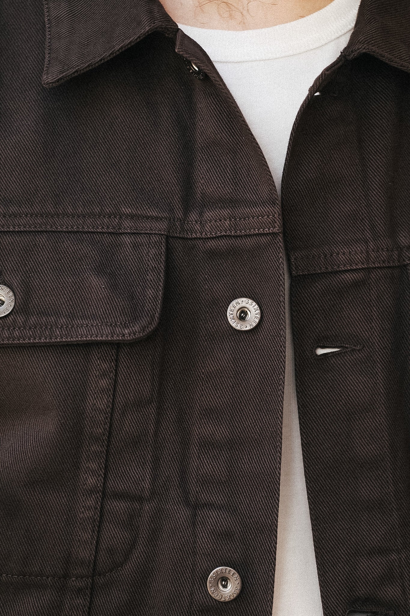 Close-up of a dark brown canvas utility jacket worn over a white t-shirt, showing the chest pocket, button details, and fabric texture.