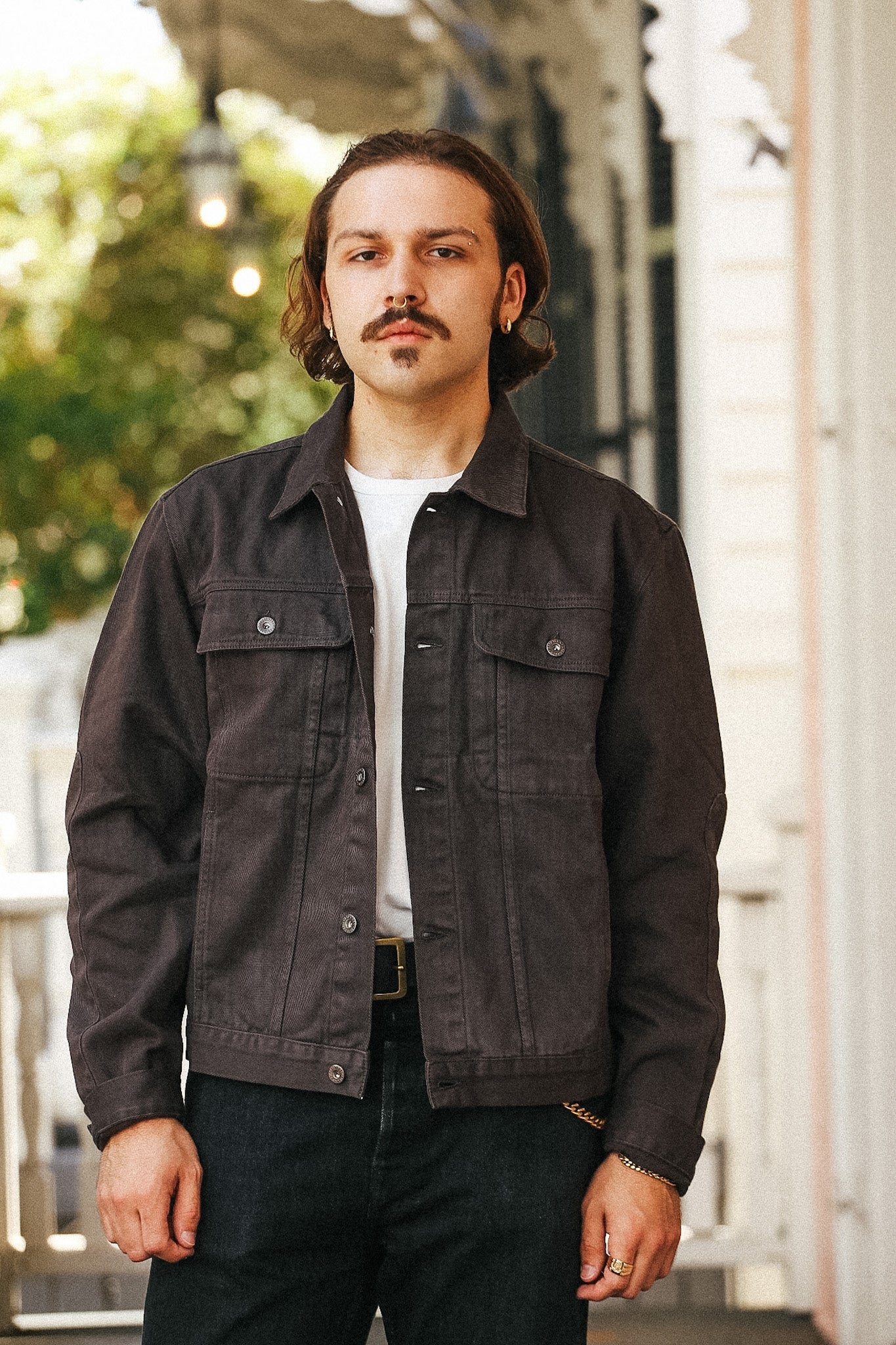 A man wearing a dark charcoal gray denim trucker jacket over a white t-shirt and dark jeans, standing on a porch with a blurred background.