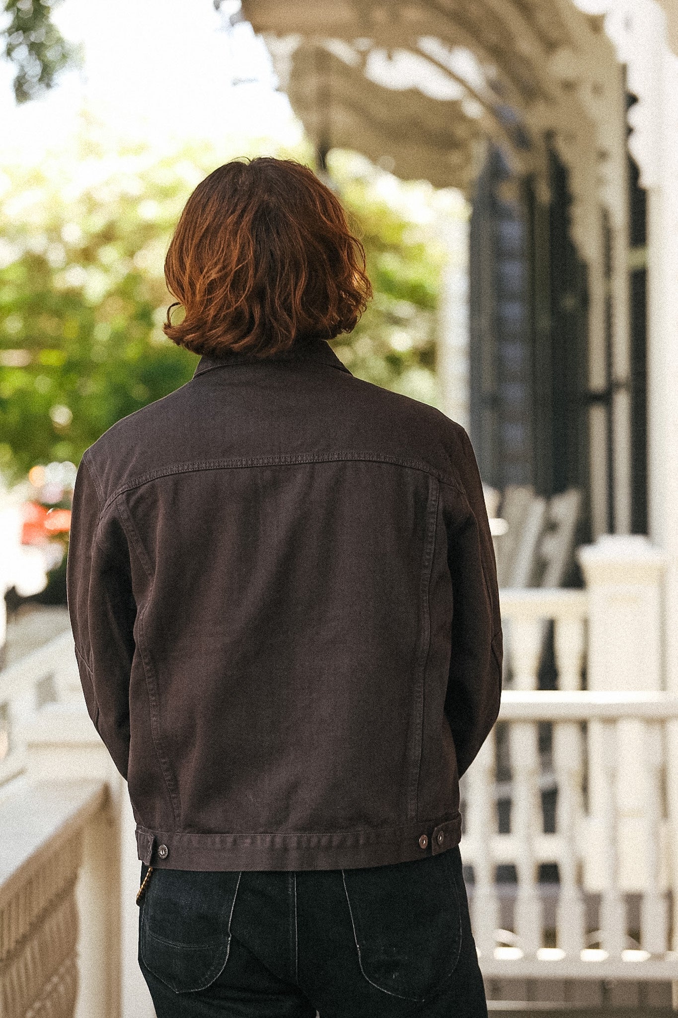 A person viewed from behind wearing a dark brown corduroy jacket and dark jeans, standing on a porch with a blurred outdoor background.