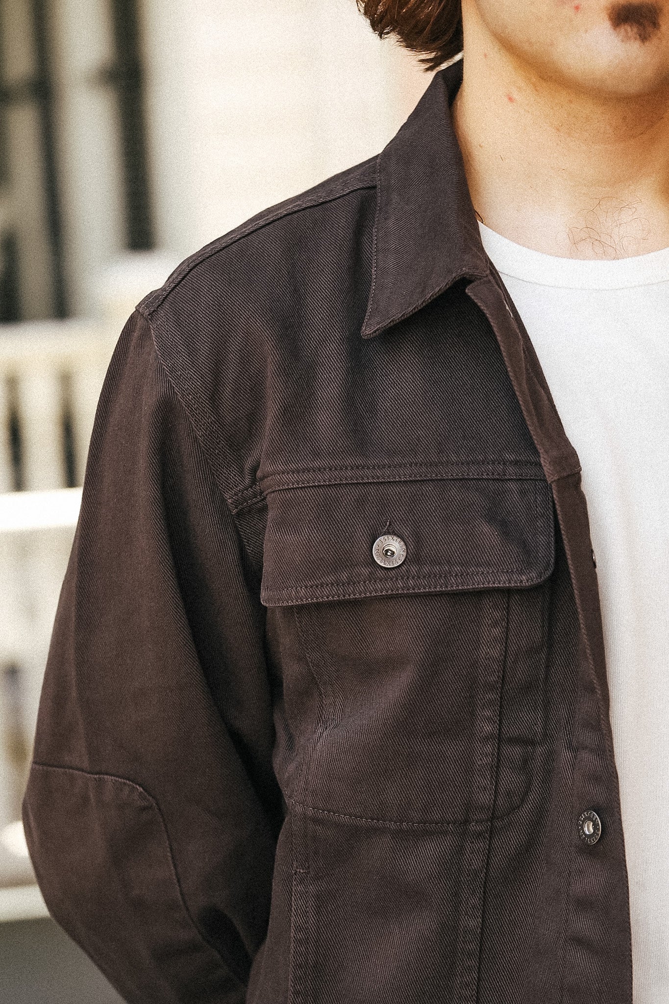 A close-up of a person wearing a dark brown canvas utility jacket over a white t-shirt, showing the chest pocket and collar.