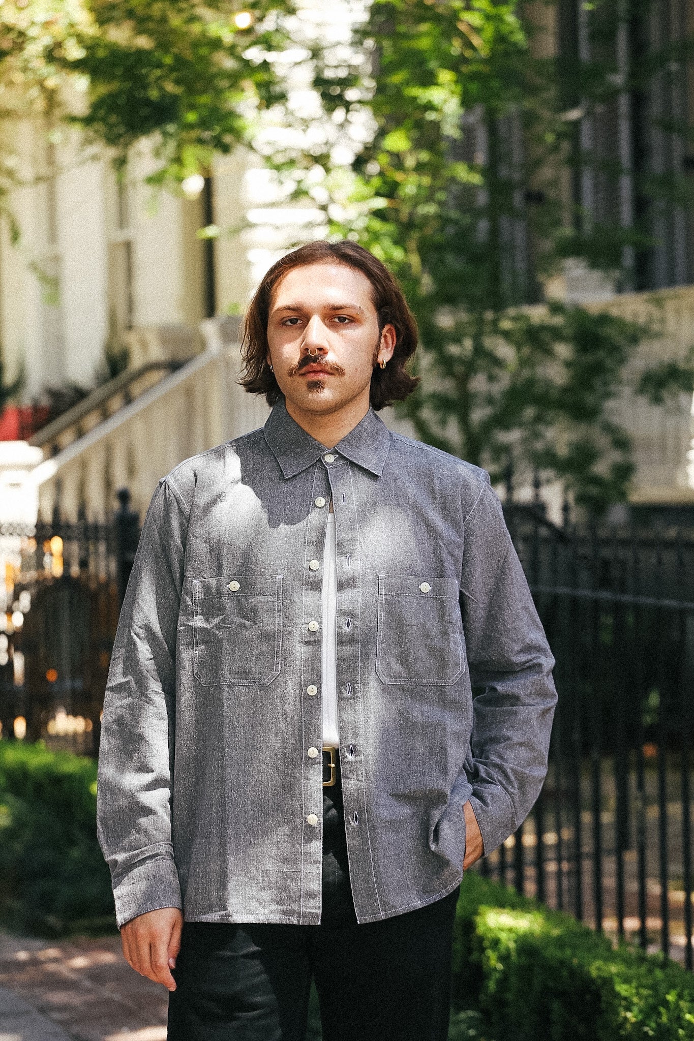 A man wearing a gray textured button-up shirt over a white undershirt and black pants, standing outdoors in front of a building with green trees and a black metal fence.