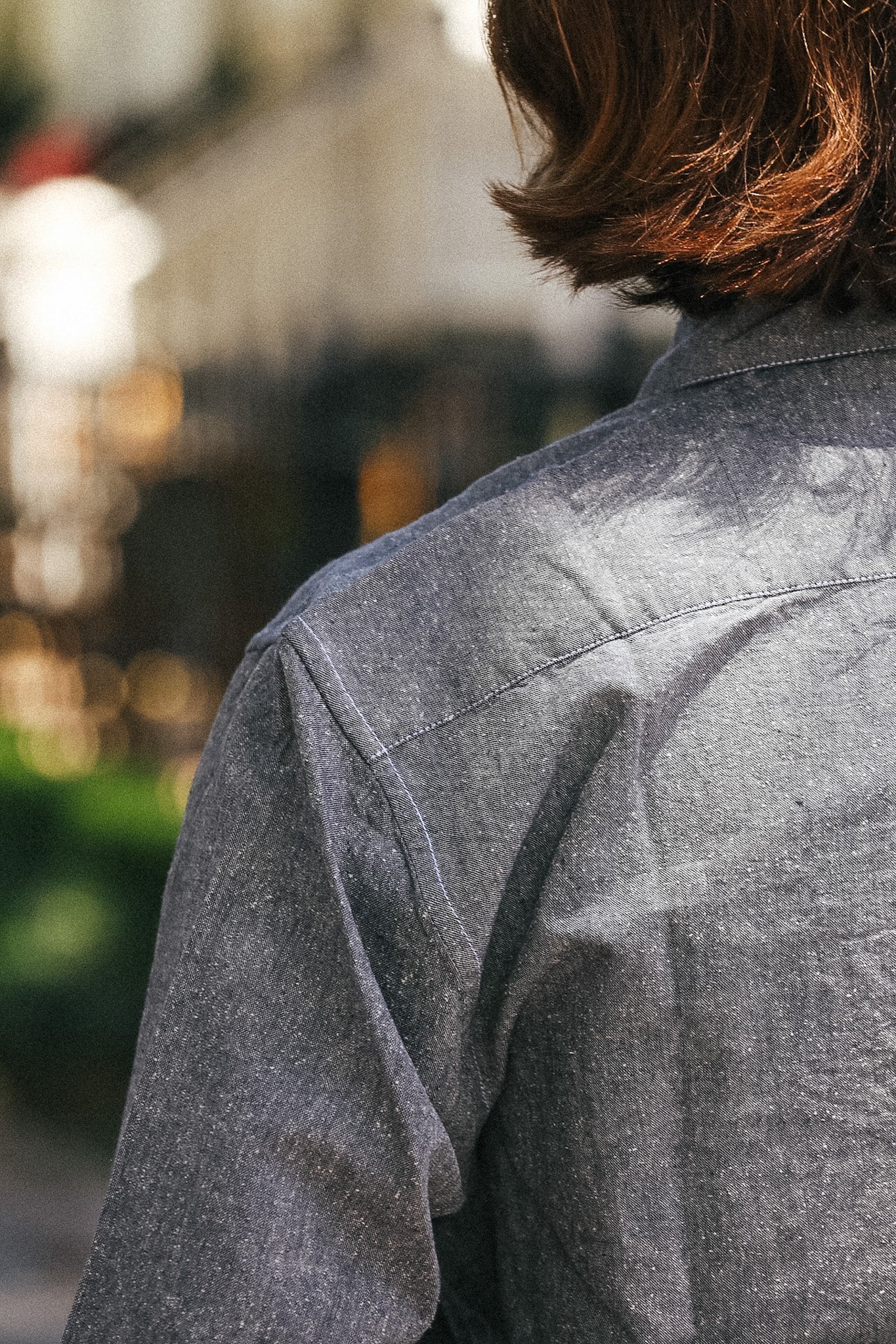 Close-up back view of a person wearing a textured gray button-up shirt in an outdoor setting with a blurred background.