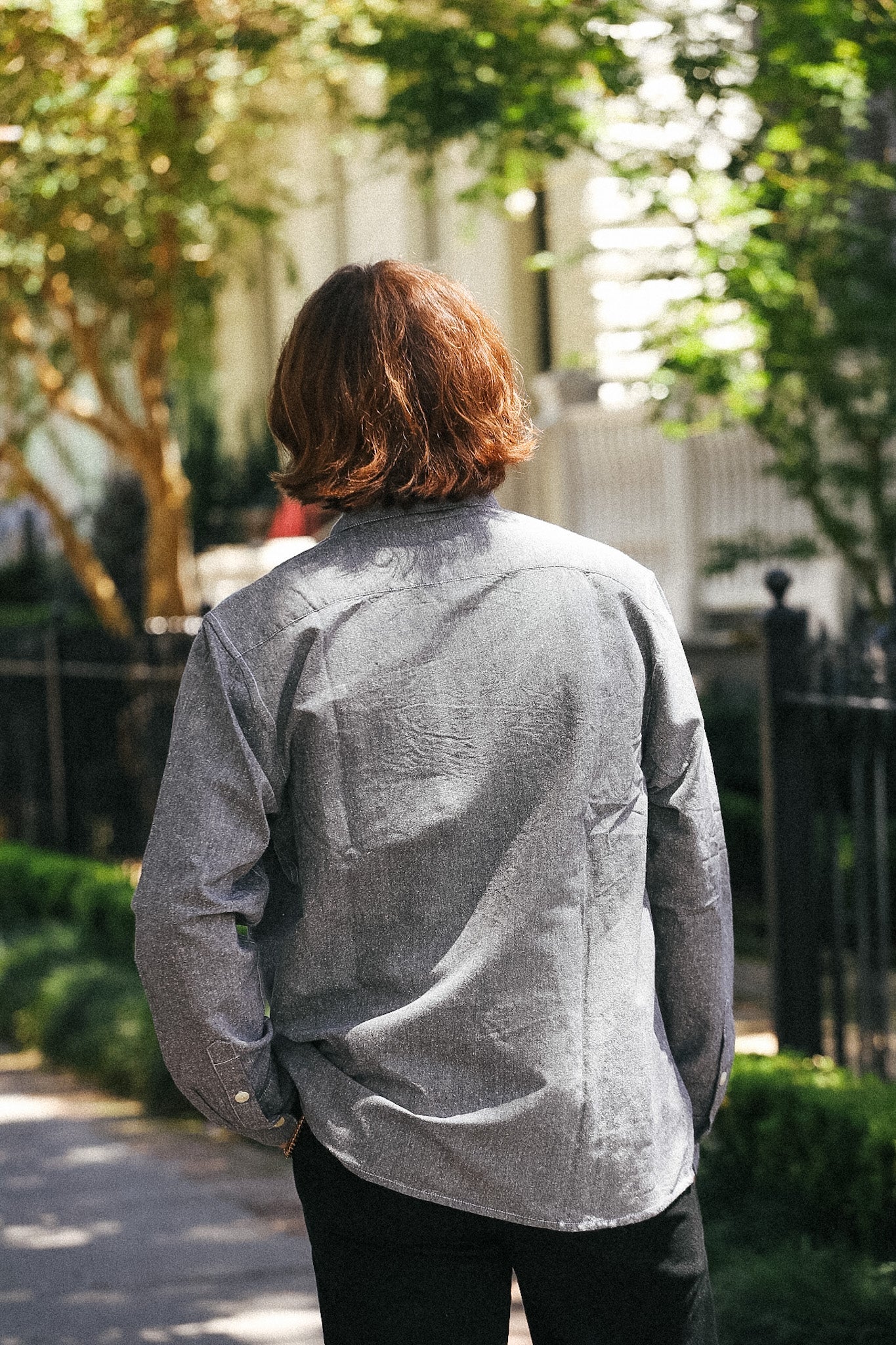 A person viewed from behind wearing a light gray textured long-sleeve button-up shirt and black pants, standing on a sunlit outdoor path with trees and a fence in the background.