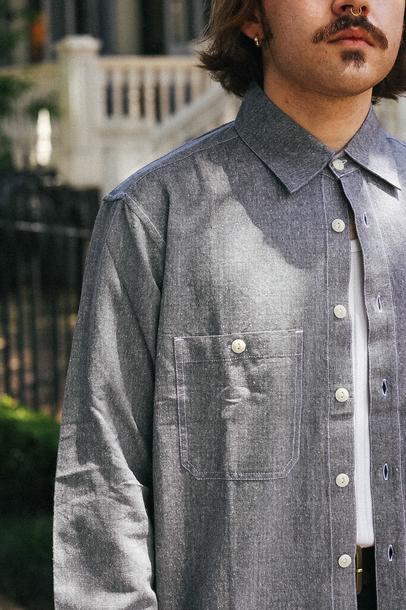 A close-up of a man wearing a textured gray long-sleeve button-up shirt with a chest pocket, layered over a white undershirt, set against an outdoor background with soft focus.