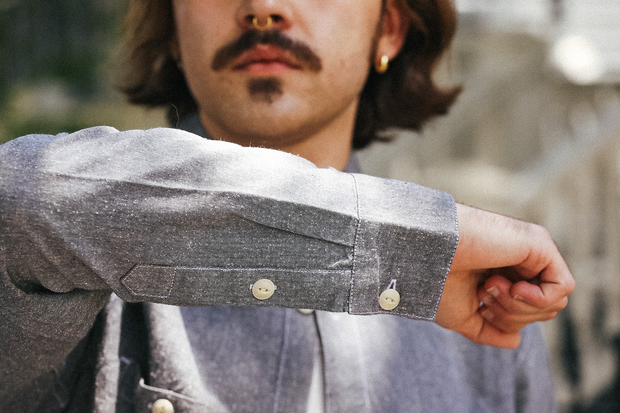 Close-up of a person wearing a textured gray long-sleeve shirt, showing the buttoned cuff and part of the chest area, with a blurred outdoor background.