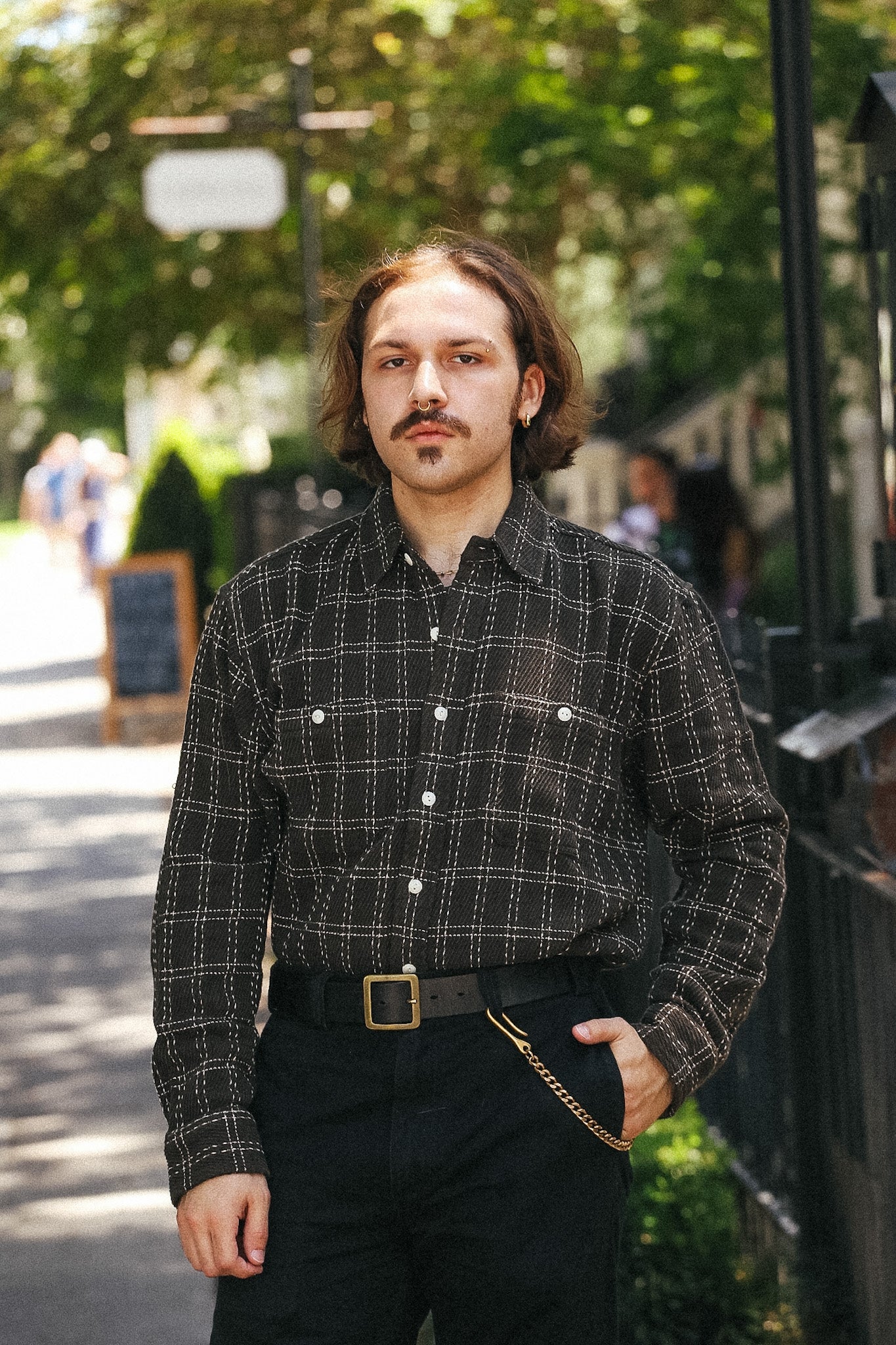 A man wearing a black and brown plaid button-down shirt with a gold chain detail at the waist, standing outdoors on a sunny day.