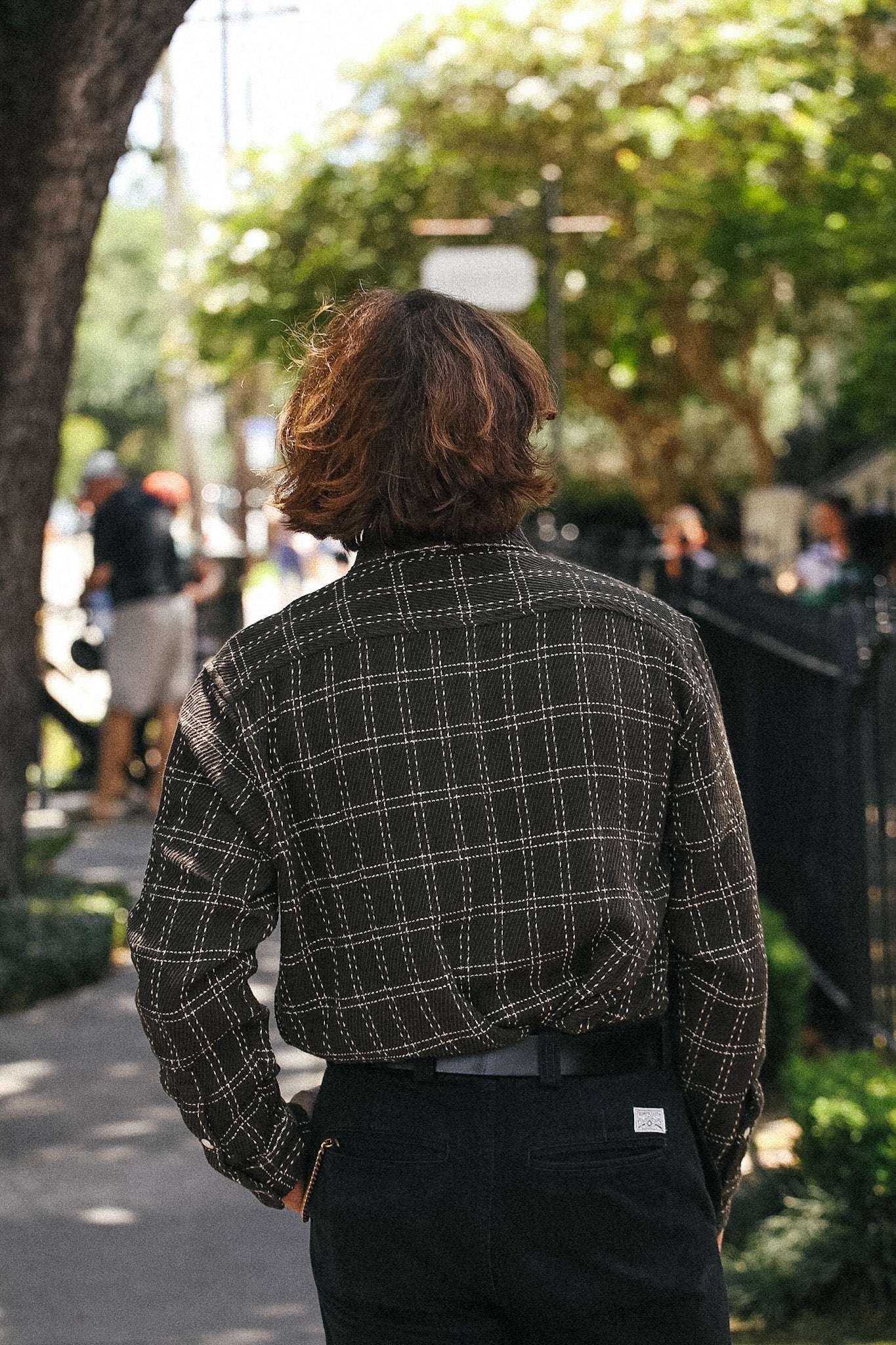 A person viewed from behind wearing a dark brown and black plaid flannel shirt and dark trousers, standing on a tree-lined sidewalk.
