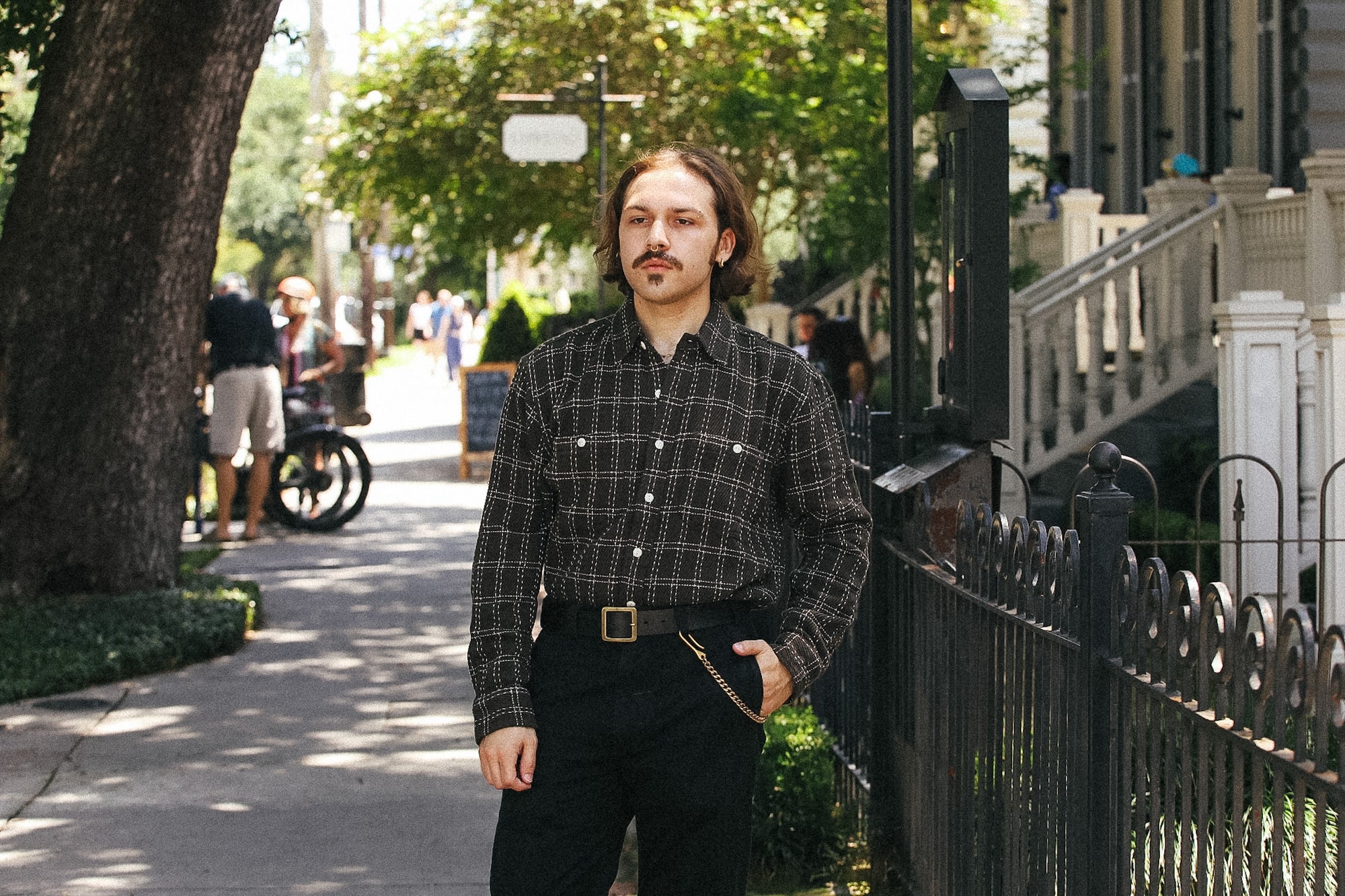 A man stands on a sunlit sidewalk wearing a dark plaid button-up shirt and black pants, with trees, a house, and a fence in the background.