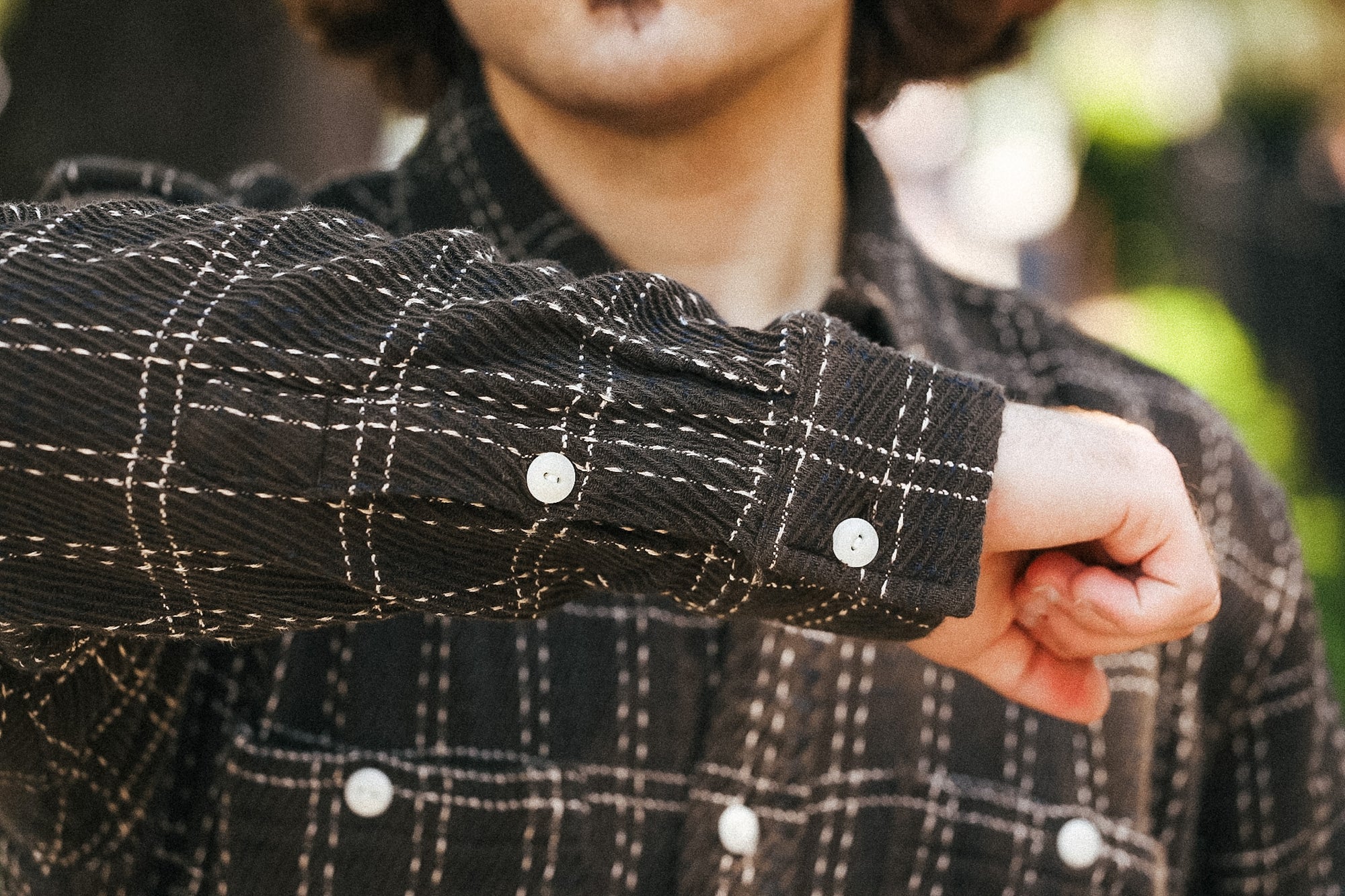 Close-up of a person wearing a dark plaid button-up shirt, focusing on the sleeve cuff with white buttons and contrast stitching.