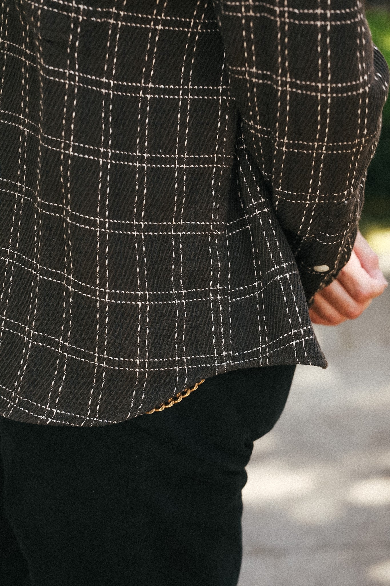 Close-up of a person wearing a dark brown plaid overshirt with white stitched windowpane lines and black pants, standing outdoors.