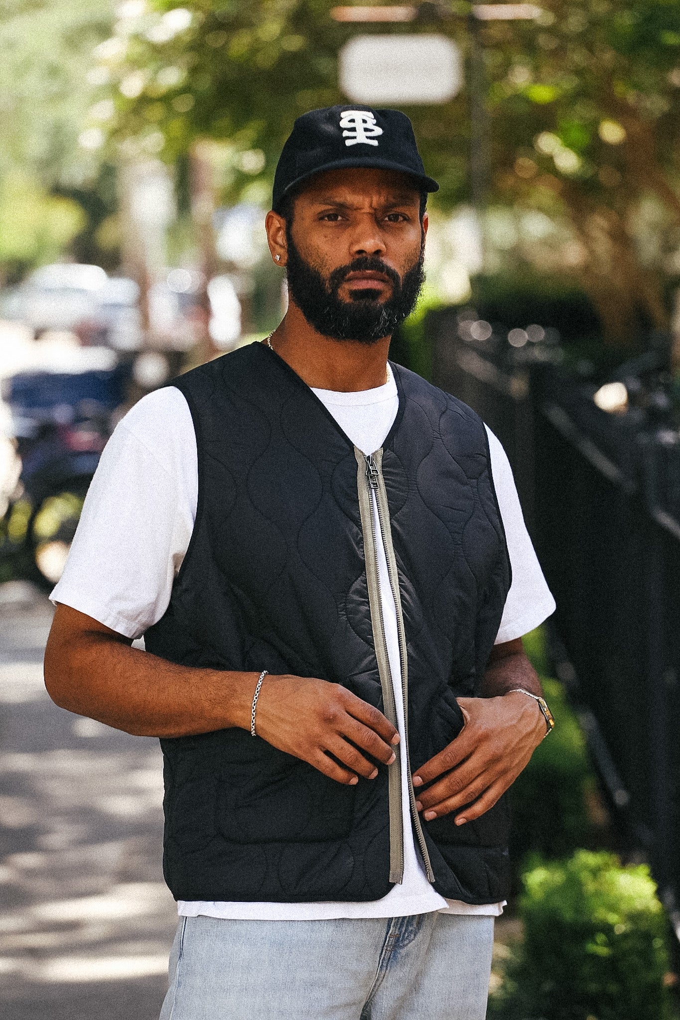 A man wearing a black quilted zip-up vest over a white t-shirt, light blue jeans, and a black baseball cap with a white logo, standing outdoors on a blurred street background.