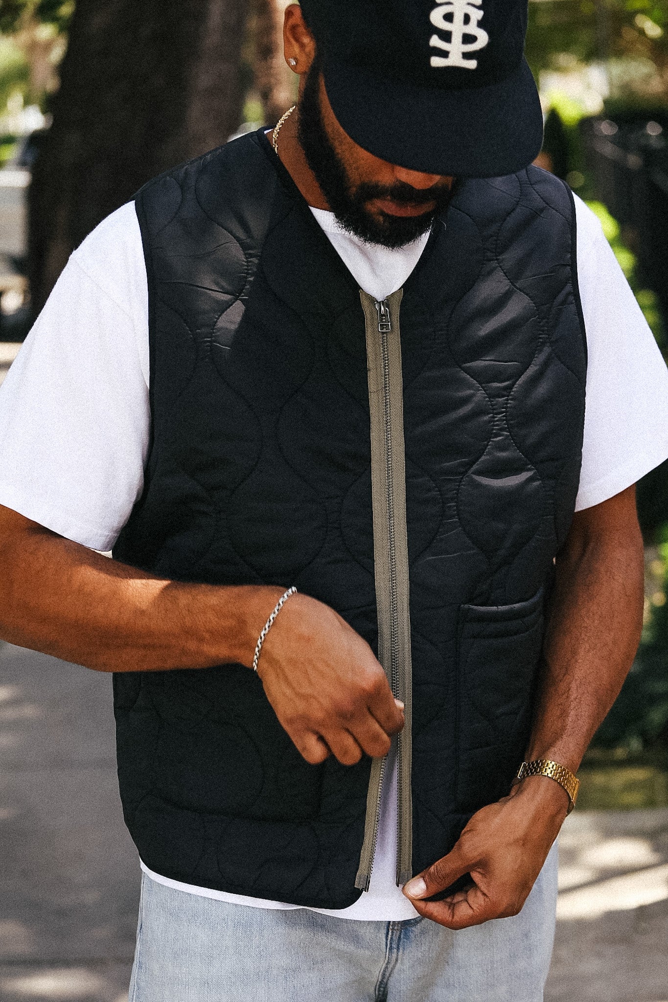 A man wearing a black quilted zip-up puffer vest over a white t-shirt and light blue jeans, standing outdoors in a park-like setting.
