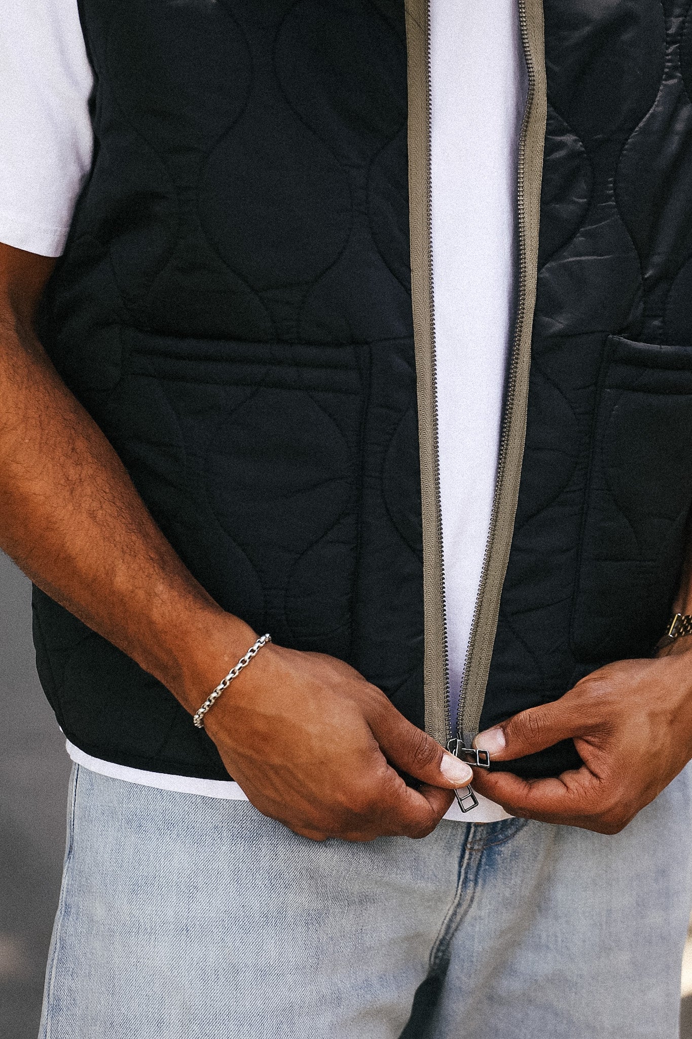 A close-up of a person wearing a black quilted zip-front vest over a white t-shirt and light blue jeans, with both hands adjusting the zipper.