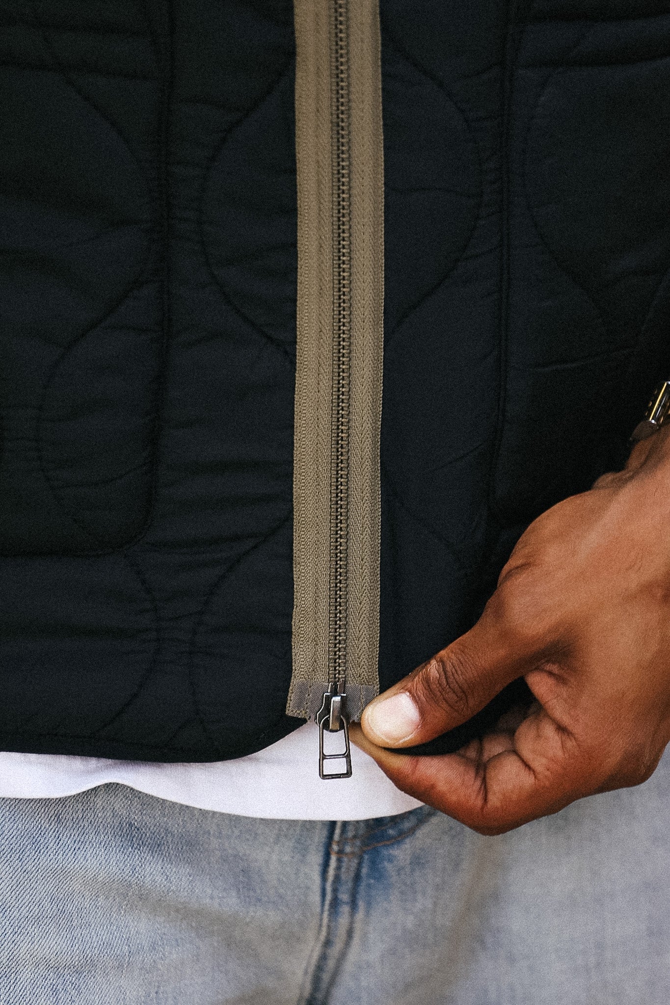 Close-up of a black quilted puffer jacket being zipped, with a hand pulling the zipper tab. The jacket has a contrasting olive green zipper track and is worn over a white shirt and light blue jeans.