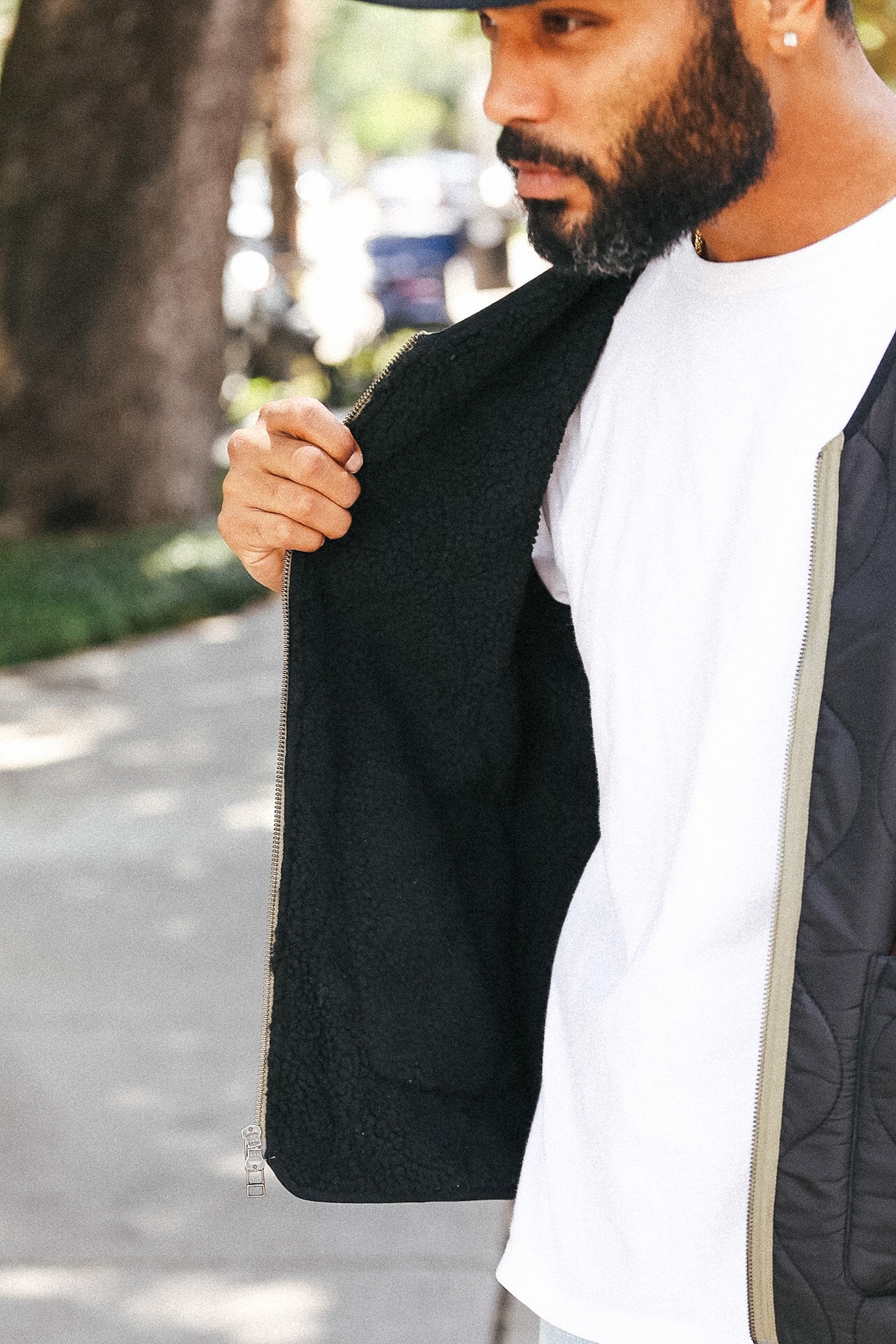A man wearing a black zip-up jacket with a plush sherpa fleece interior, shown in an outdoor setting next to a white t-shirt.