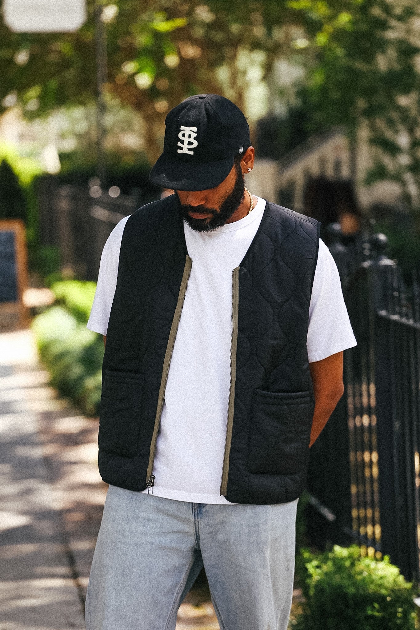 A man wearing a black quilted puffer vest over a white t-shirt, light gray jeans, and a black baseball cap with a white monogram logo, standing on an outdoor sidewalk.