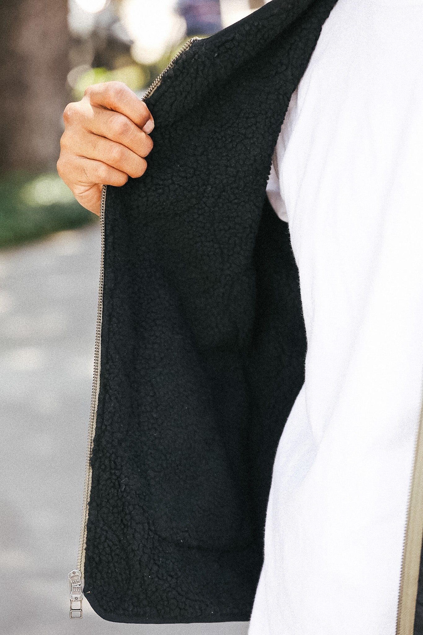 Close-up of a person wearing a black sherpa-lined zip jacket over a white shirt, holding the zipper open with a hand outdoors.