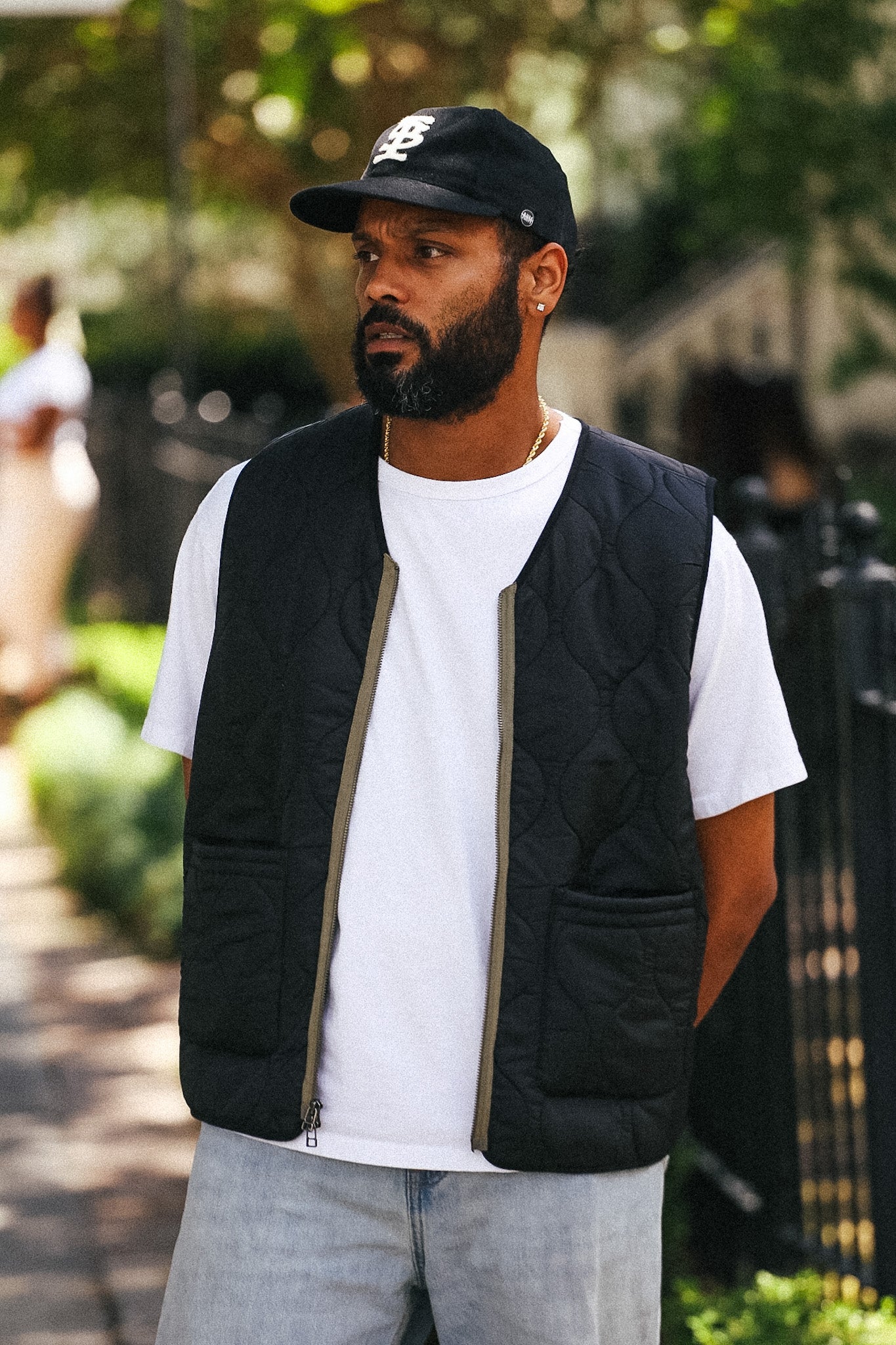 A man wearing a black quilted zip-up vest over a white t-shirt, light gray jeans, and a black baseball cap, standing outdoors on a sunny day.