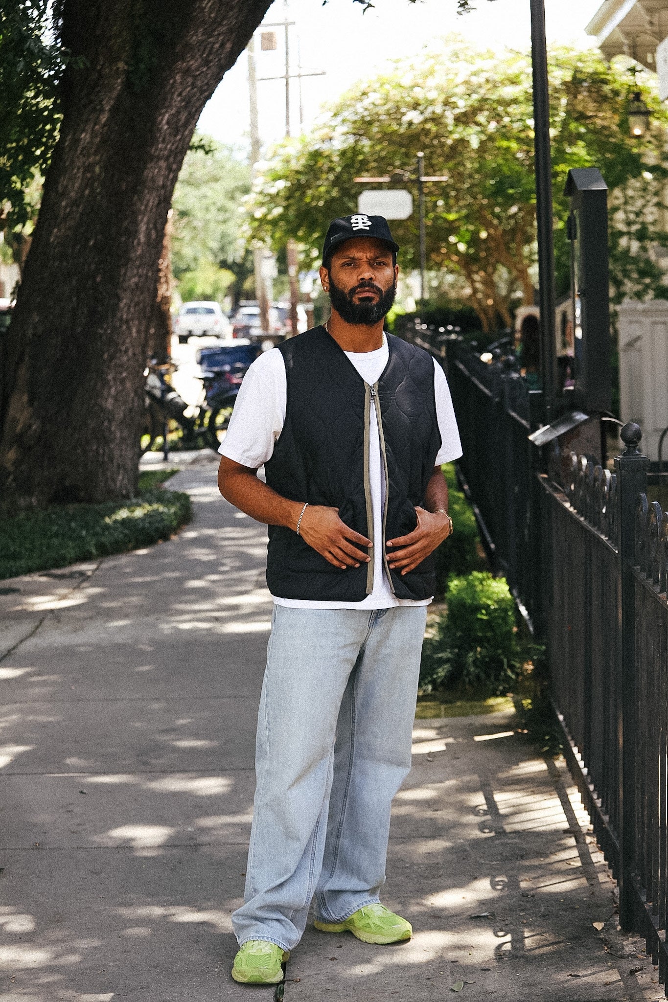 A man stands on a sunny sidewalk wearing a black zip-up vest over a white t-shirt, light-wash jeans, and neon green sneakers, with a black baseball cap. Trees, a fence, and parked cars are visible in the background.