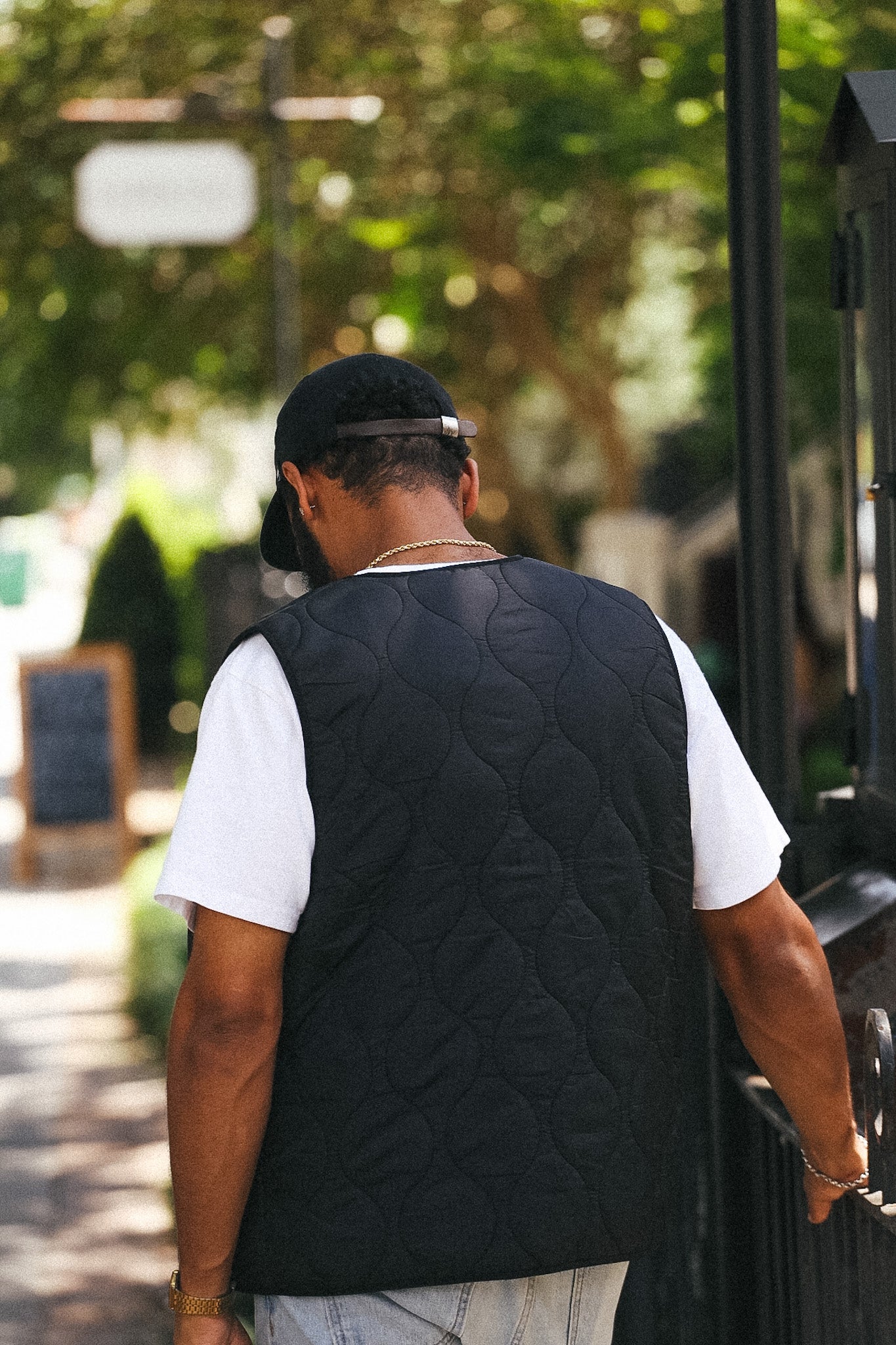A man viewed from behind wearing a black quilted vest over a white t-shirt and a black baseball cap, walking outdoors on a sunny day.