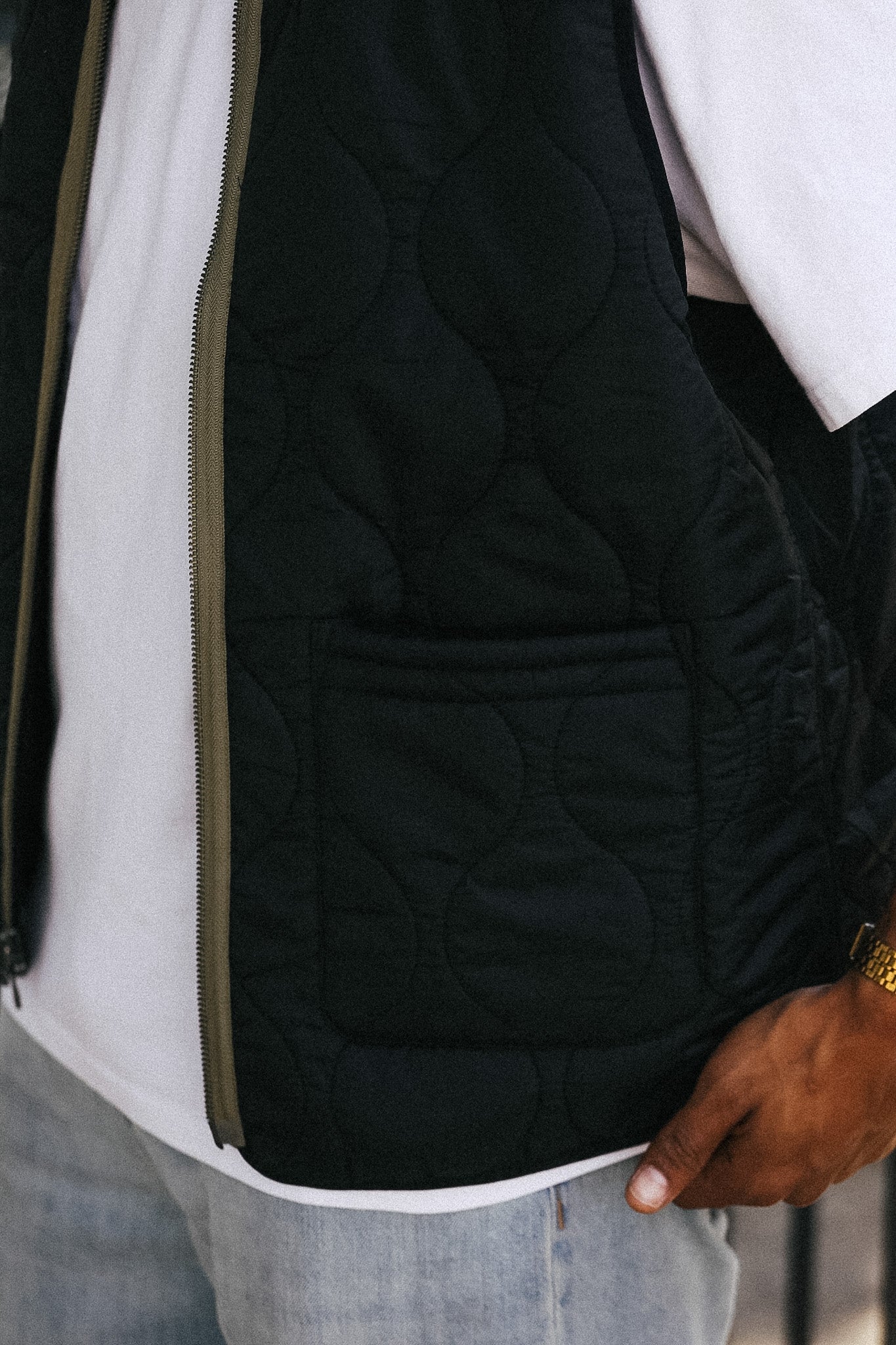 Close-up of a person wearing a black quilted puffer vest over a white t-shirt and light blue jeans, with a hand resting near the hem.