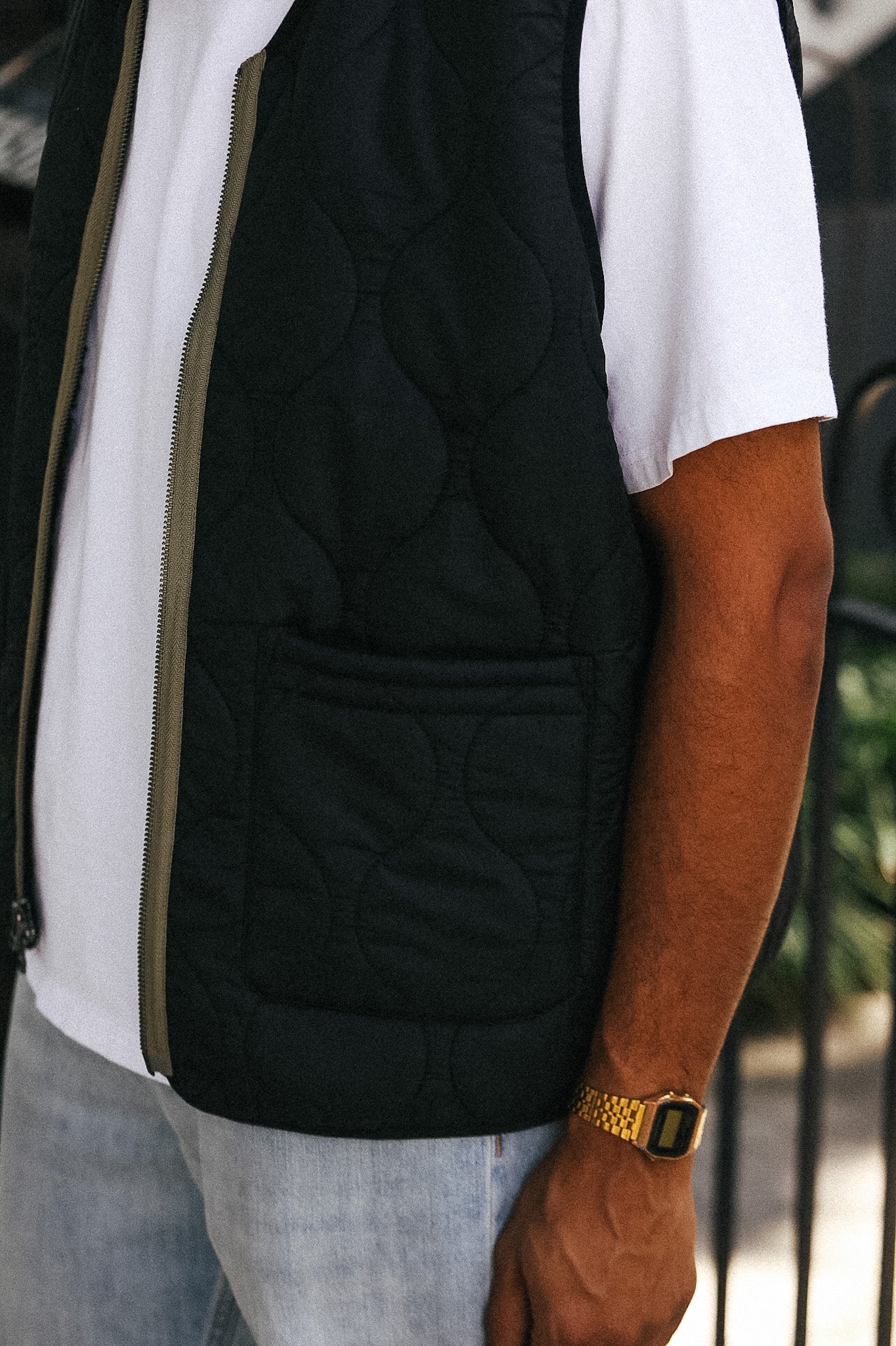 A close-up of a person wearing a black quilted zip-front vest over a white t-shirt and light blue jeans, with a gold watch visible on the wrist.