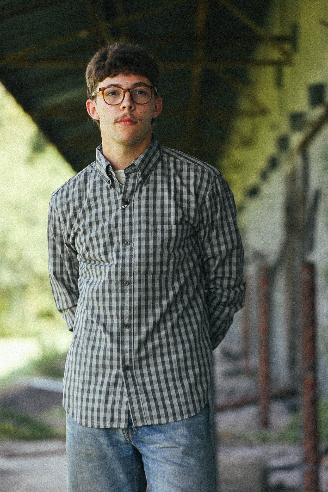 A young man with glasses and a mustache wearing a black and white checkered button-down shirt and blue jeans, standing under a covered walkway.