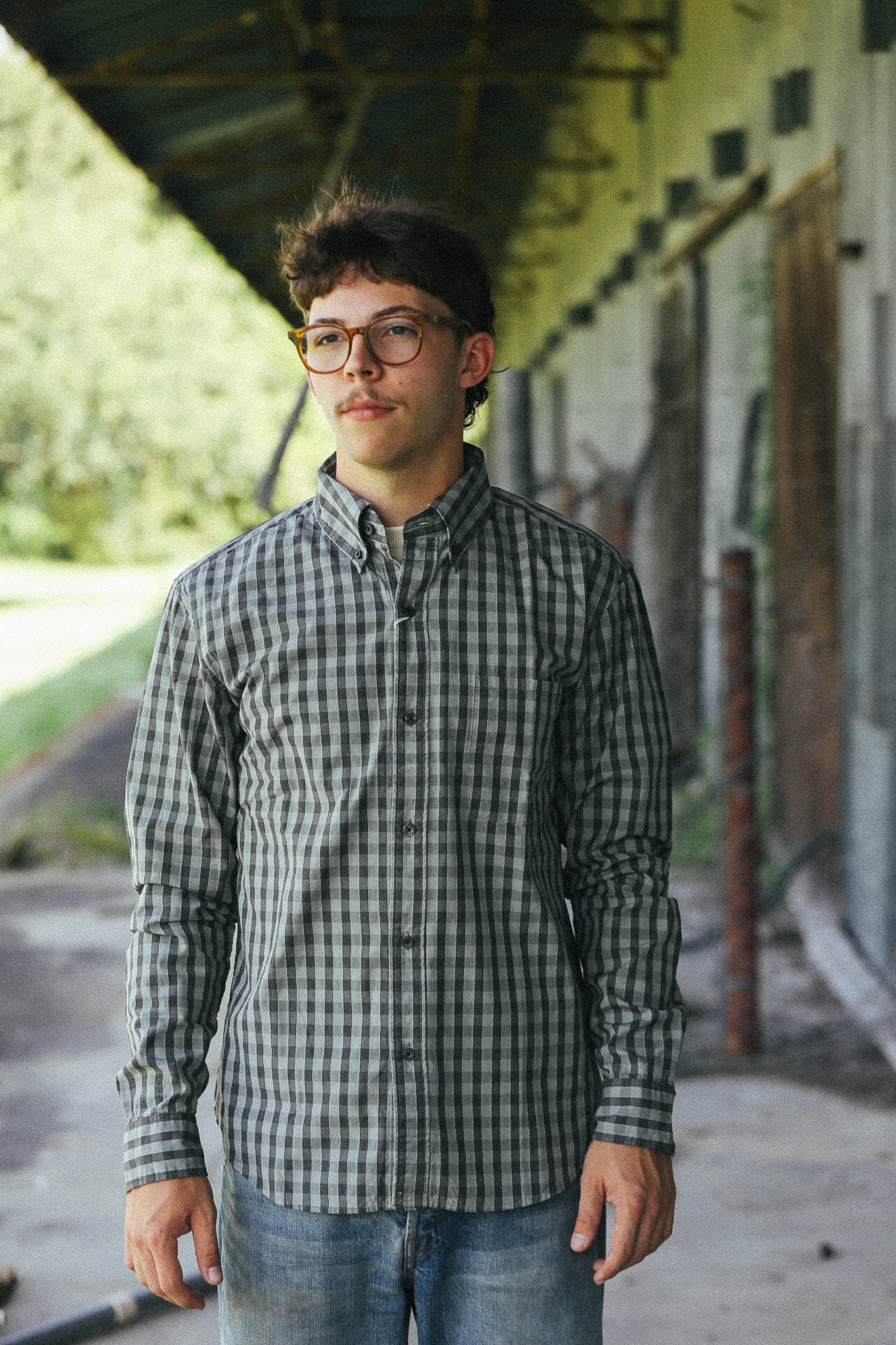 A young man with glasses stands in a covered outdoor walkway wearing a long-sleeve black and white checkered button-down shirt and blue jeans.