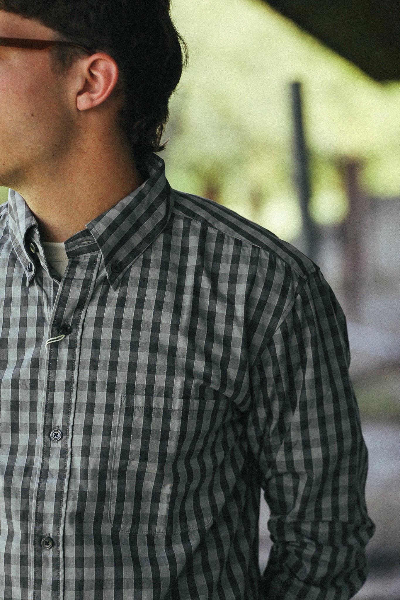 A close-up side view of a man wearing a dark gray and black checkered button-down shirt, with a blurred outdoor background.