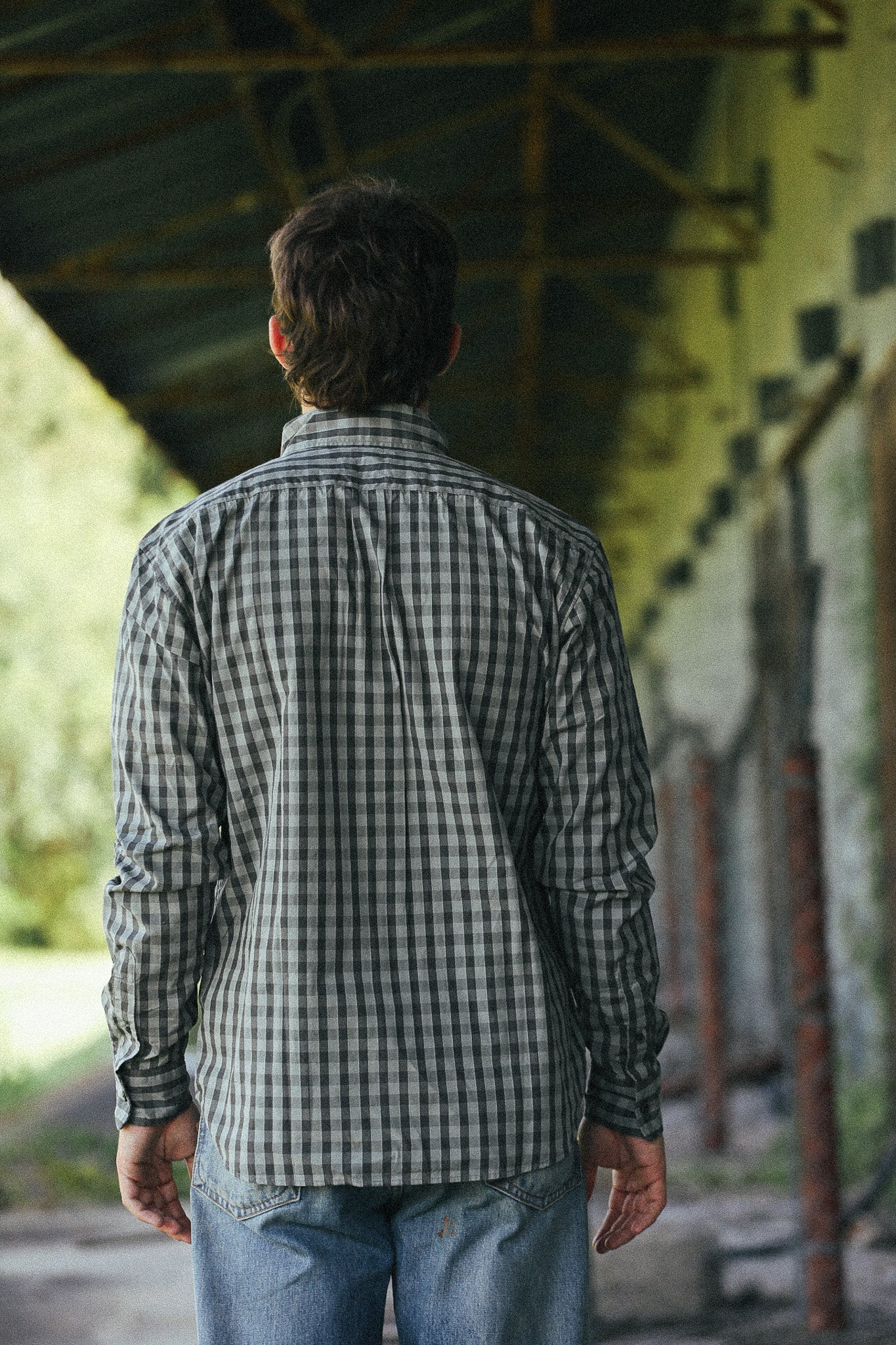 A man viewed from behind wearing a dark green and black checkered long-sleeve button-up shirt and blue jeans, standing in a covered outdoor walkway.