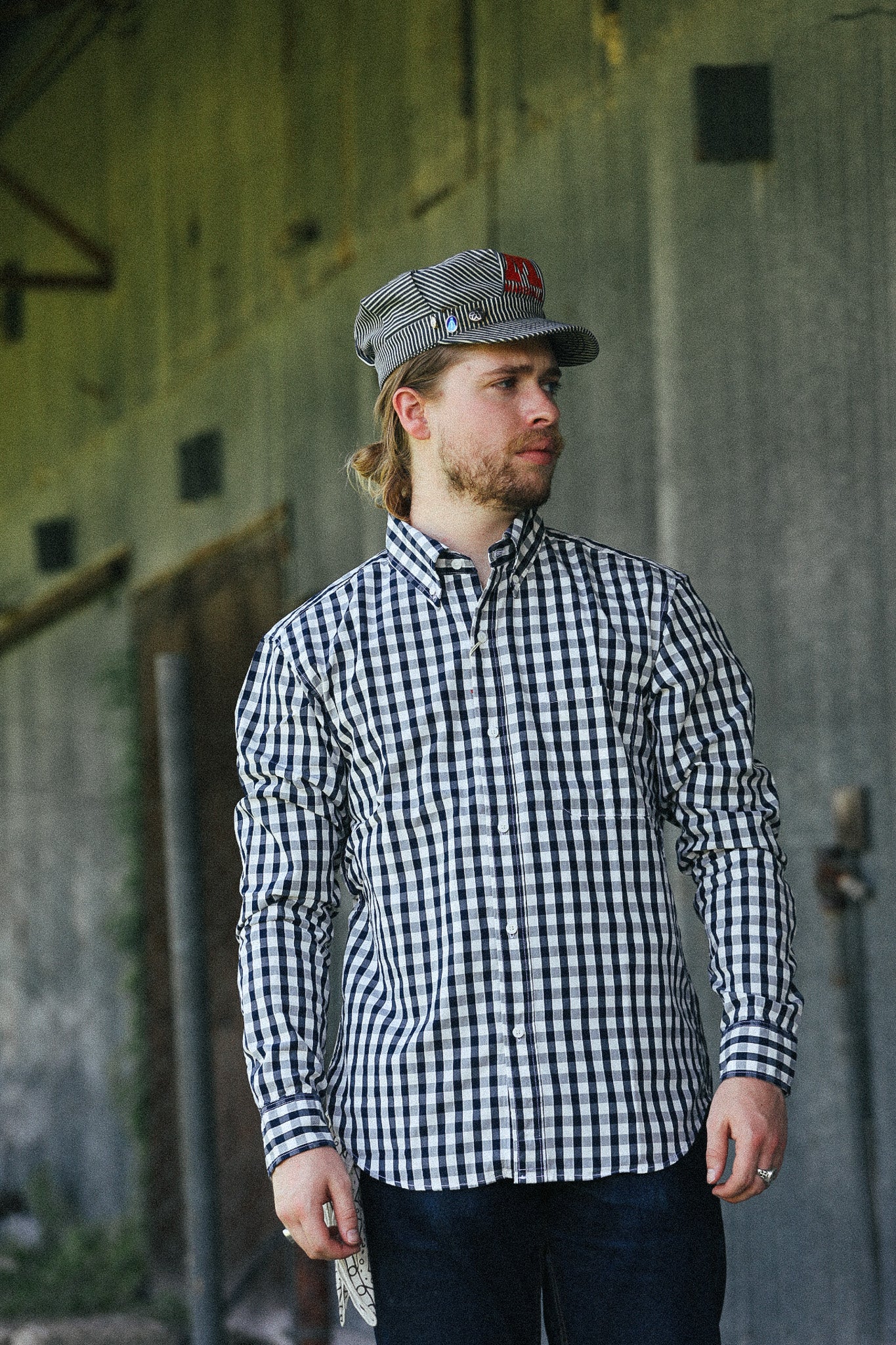 A man wearing a black and white gingham long-sleeve button-down shirt and a matching gingham cap with a red logo patch, standing in front of a rustic concrete wall.