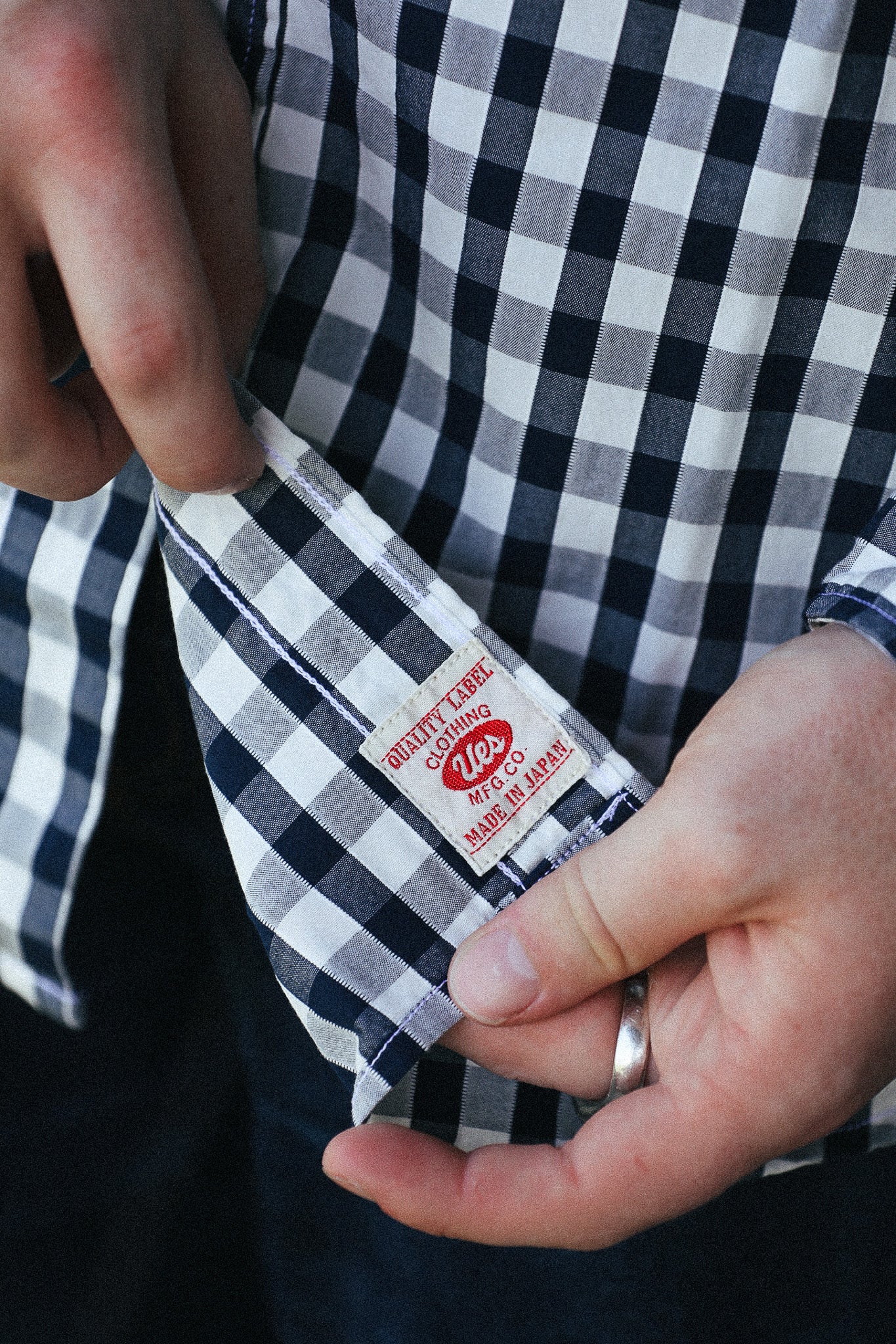 Close-up of a person's hands holding a cuff of a navy and white gingham shirt, showing the inside hem with a red and white 'Quality Cards 140 Made in Japan' label.