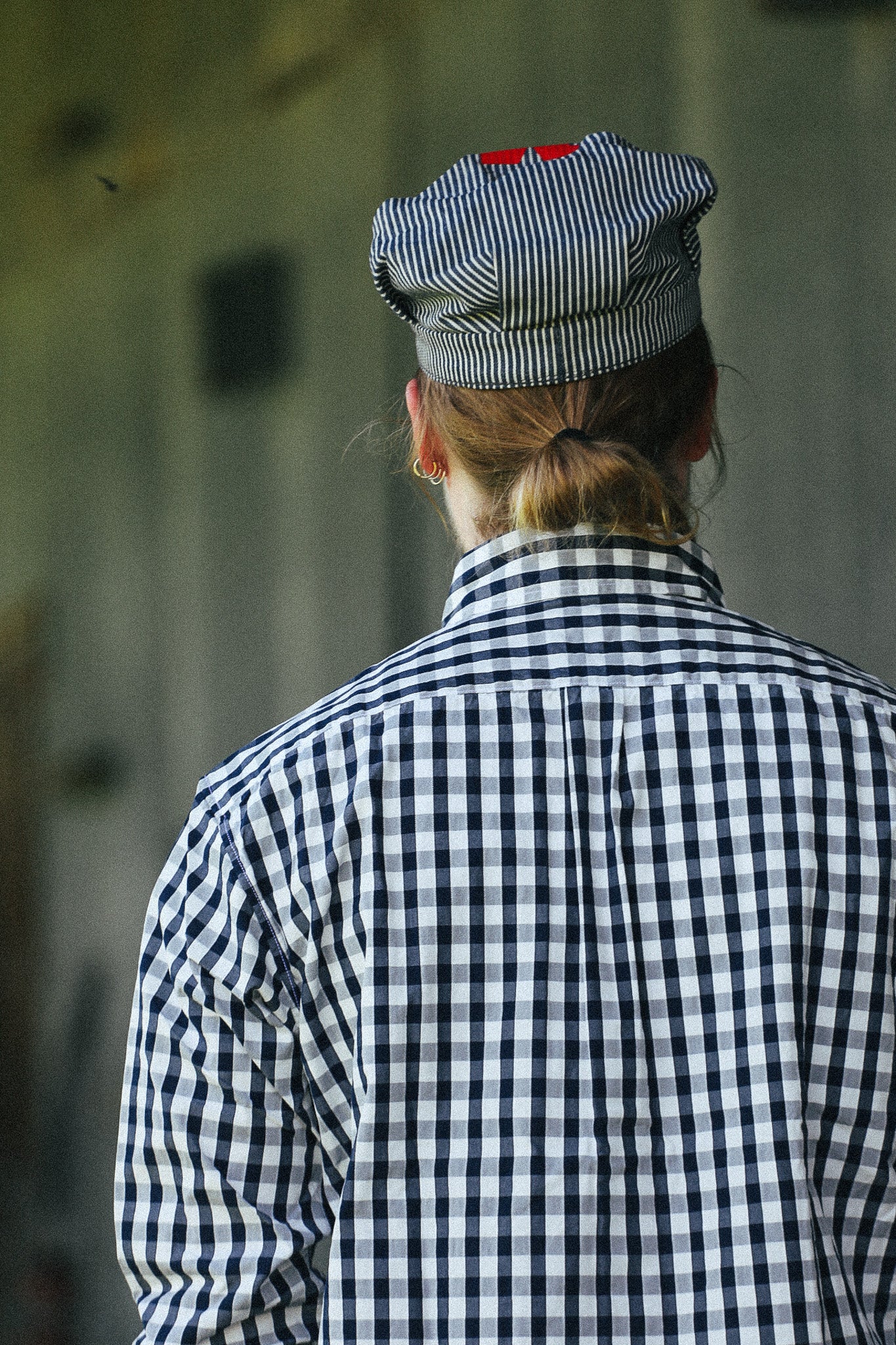 A person viewed from behind wearing a black and white gingham shirt and a matching striped cap with a small red accent, standing against a blurred gray background.