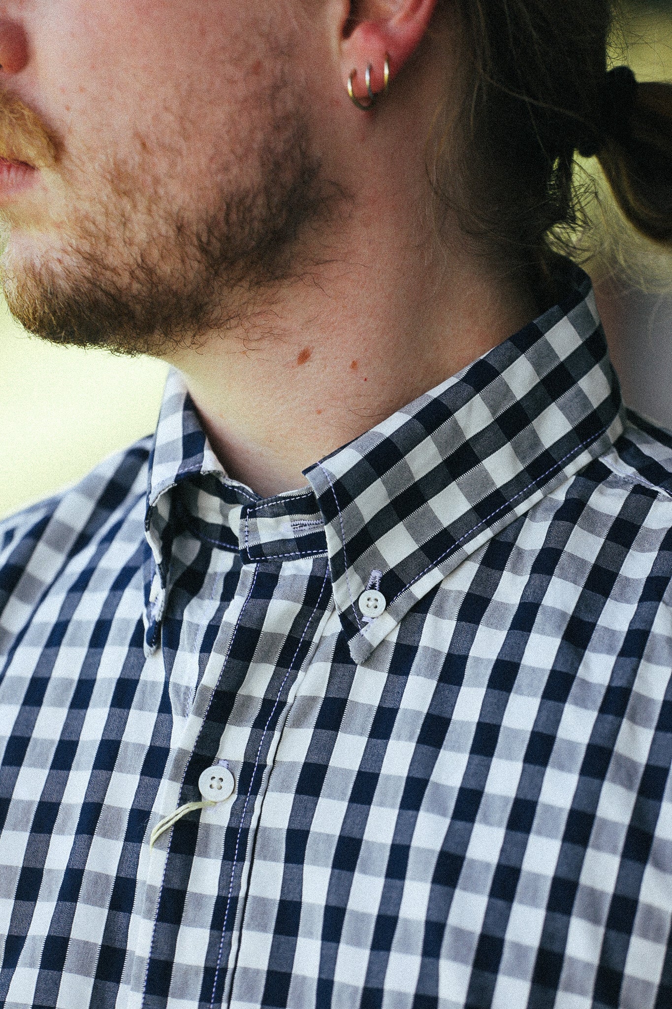 Close-up of a man wearing a black and white gingham button-down shirt, showing his neck, chin, and side profile against a softly blurred light background.