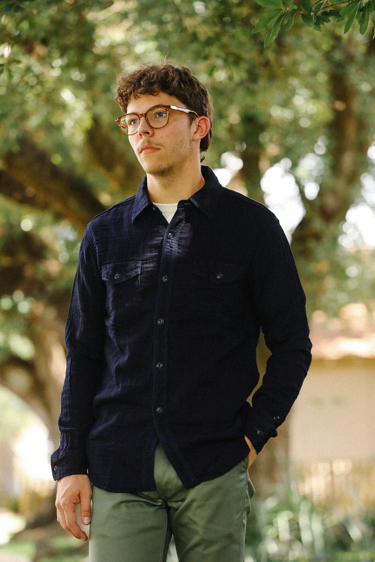 A man wearing a dark navy blue long-sleeve button-up shirt and olive green pants, standing outdoors in front of a blurred background of trees.