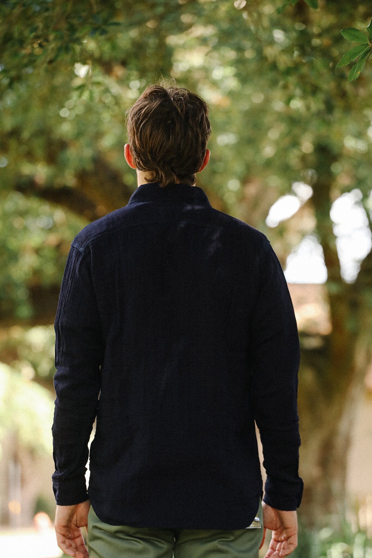 A man viewed from behind wearing a dark navy blue long-sleeve shirt and olive green pants, standing outdoors in front of a blurred background of green trees.