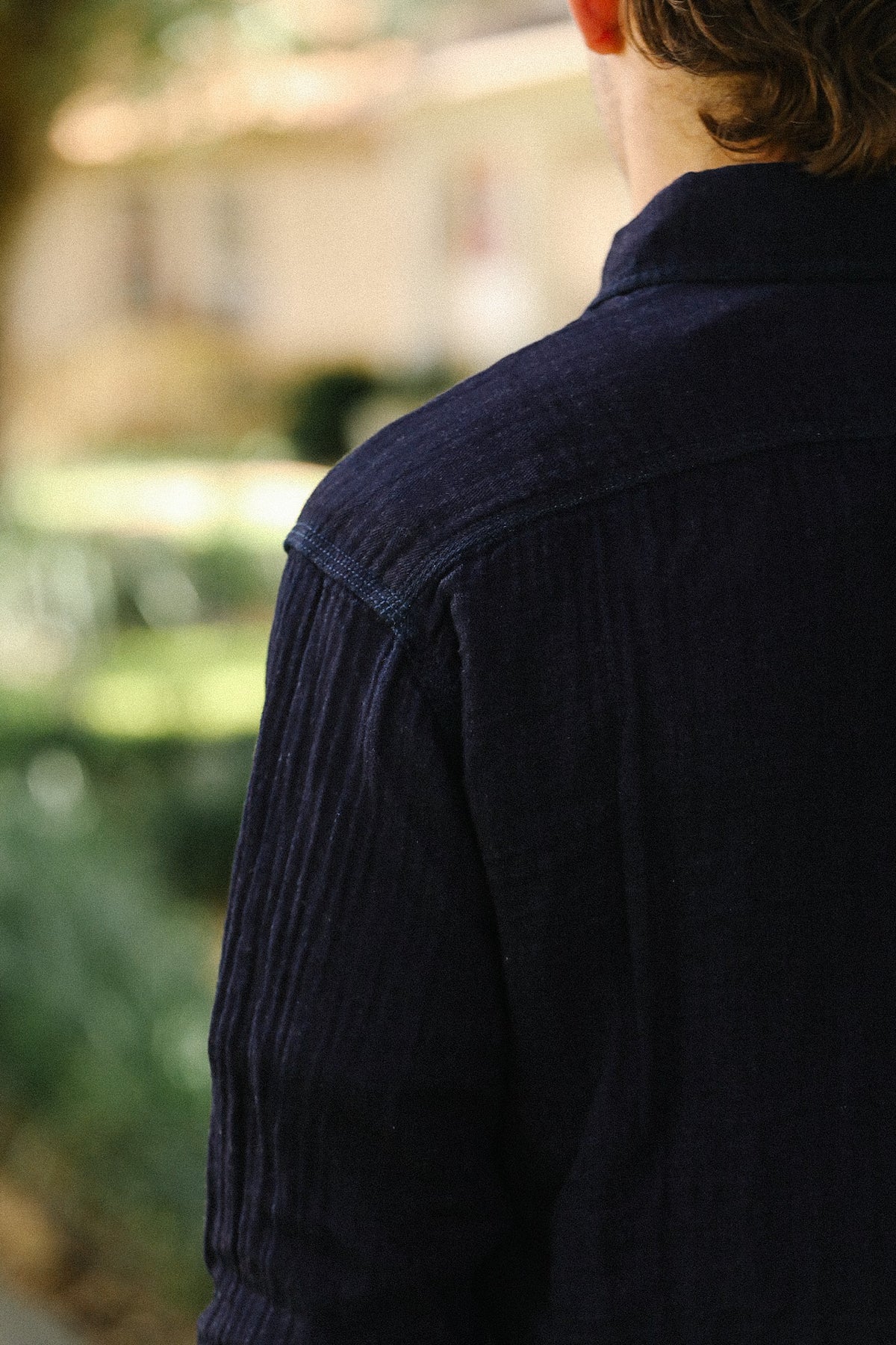 A close-up back view of a person wearing a dark navy blue corduroy shirt jacket in an outdoor setting with a blurred background.