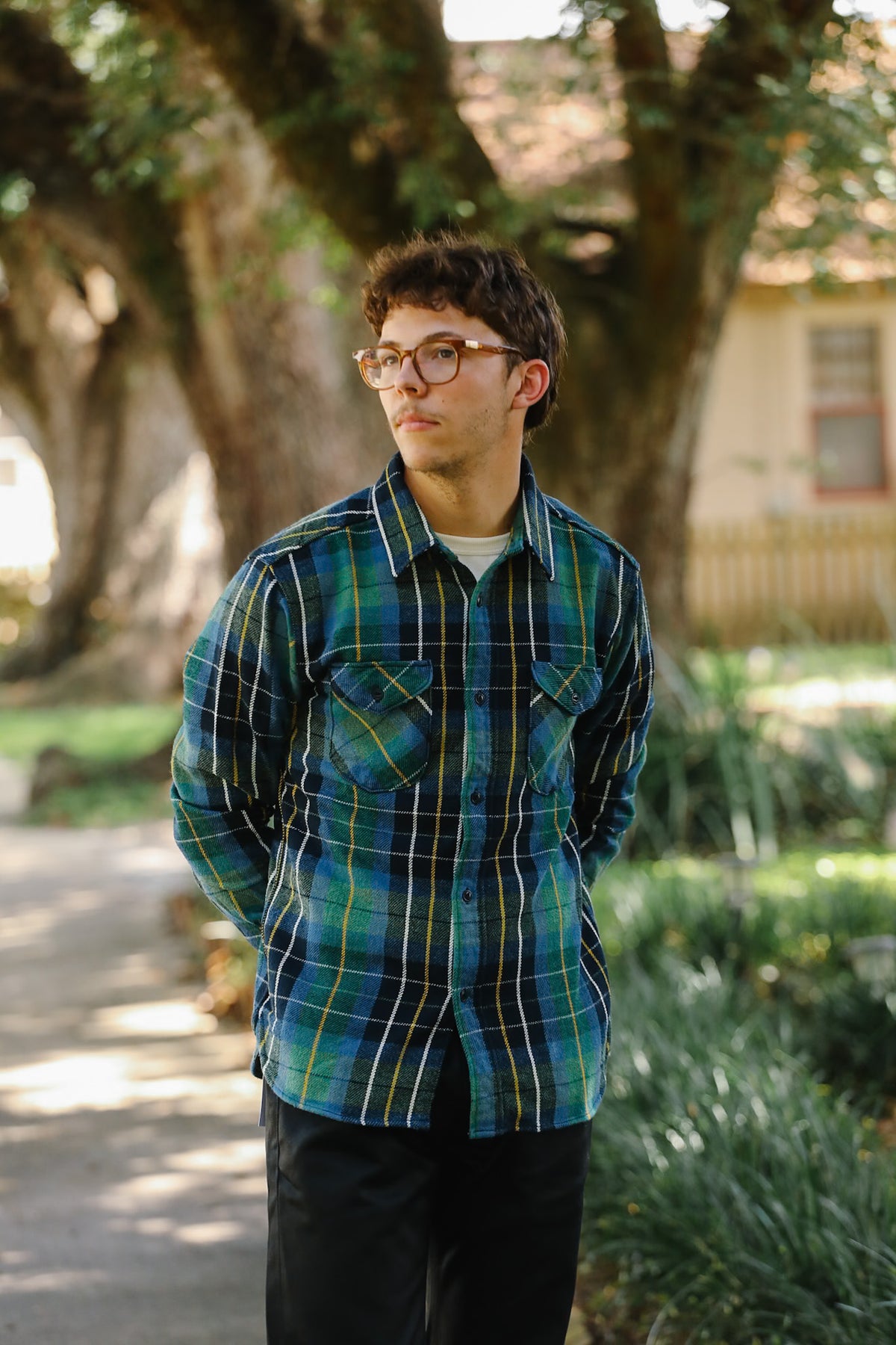A young man wearing a green and blue plaid button-down shirt with thin metal-framed glasses, standing outdoors on a paved path with trees and greenery in the background.
