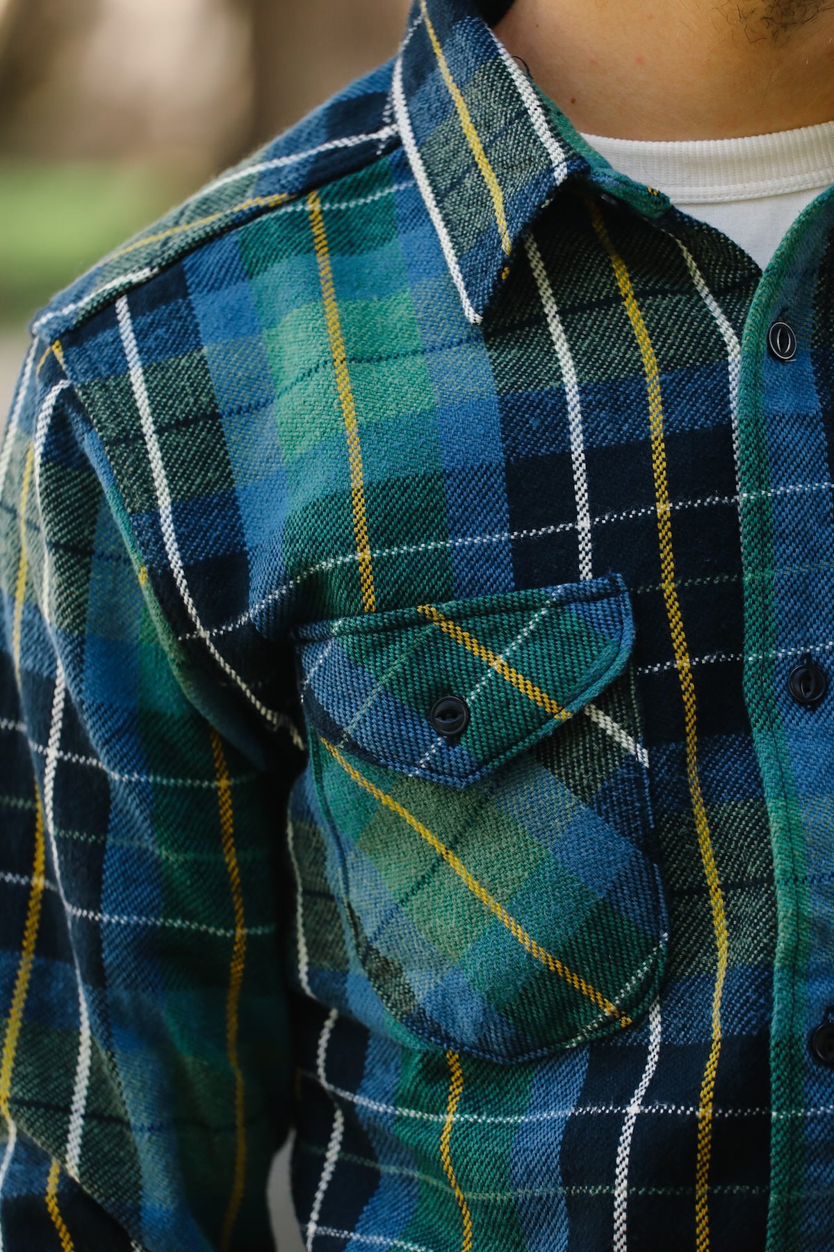 Close-up of a person wearing a blue and green plaid flannel button-up shirt with a chest pocket, over a white undershirt.
