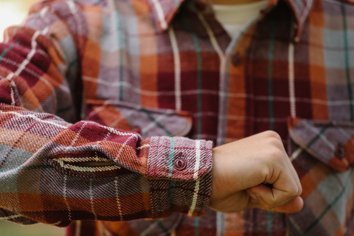 Close-up of a person wearing a long-sleeve plaid flannel shirt in rust orange, burgundy, and gray tones, with the right sleeve rolled up and the hand visible at the cuff.