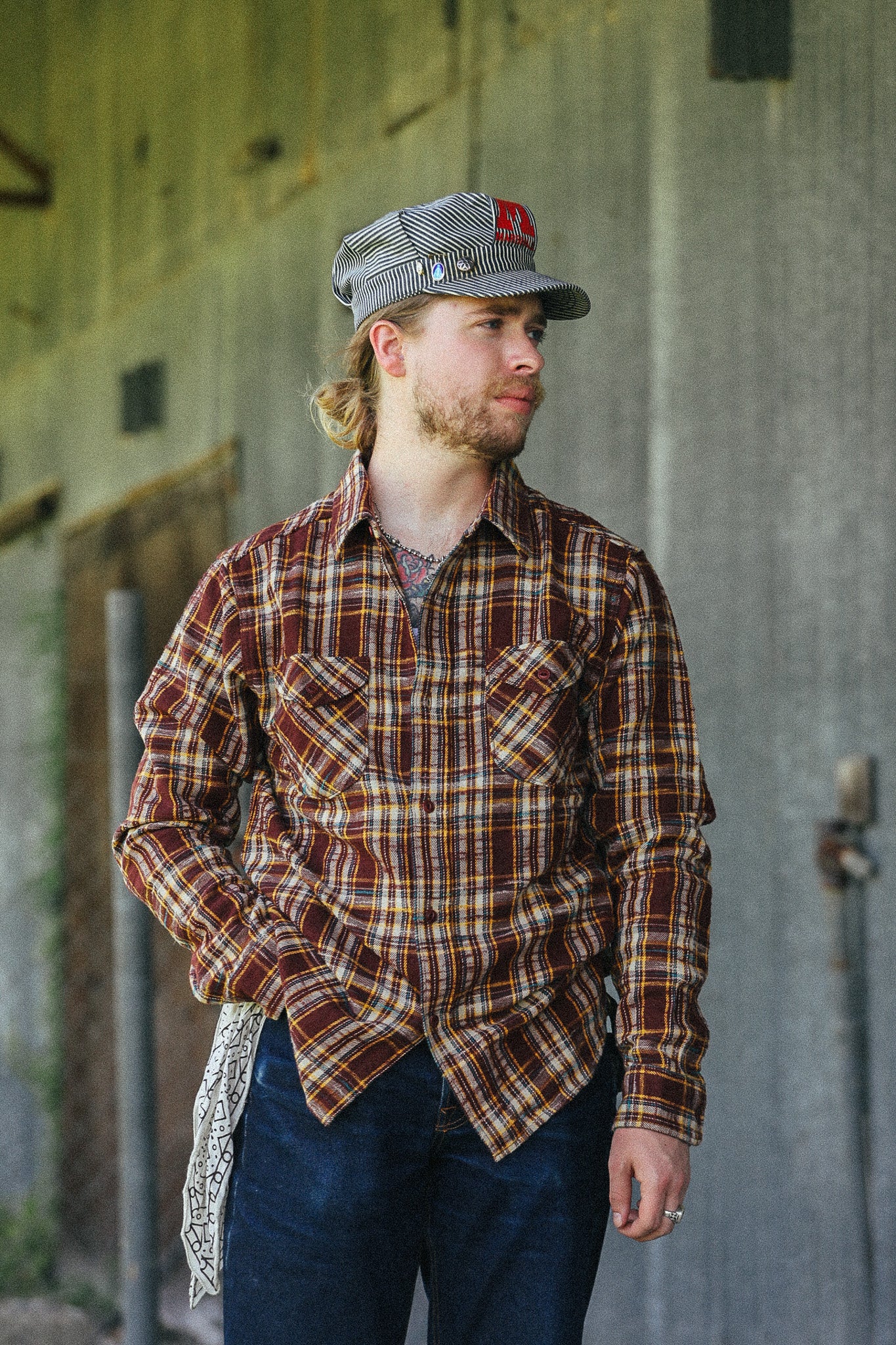 A man wearing a brown and beige plaid flannel shirt, dark blue jeans, and a gray cap with a red logo, standing in front of a rustic concrete wall.