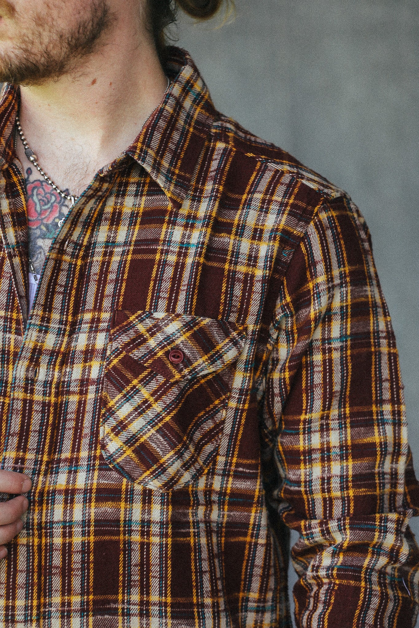 A close-up of a man wearing a brown, yellow, and black plaid button-down shirt, showing the chest pocket and collar area.