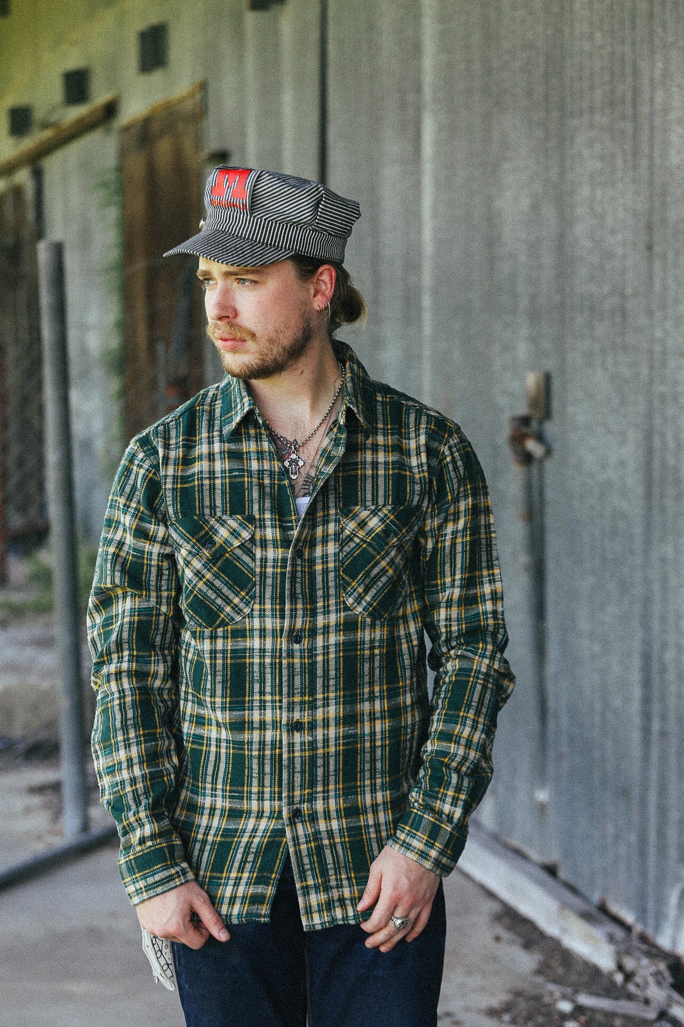 A man wearing a green and yellow plaid button-down shirt, dark jeans, and a gray striped cap with a red patch, standing in front of a corrugated metal wall.