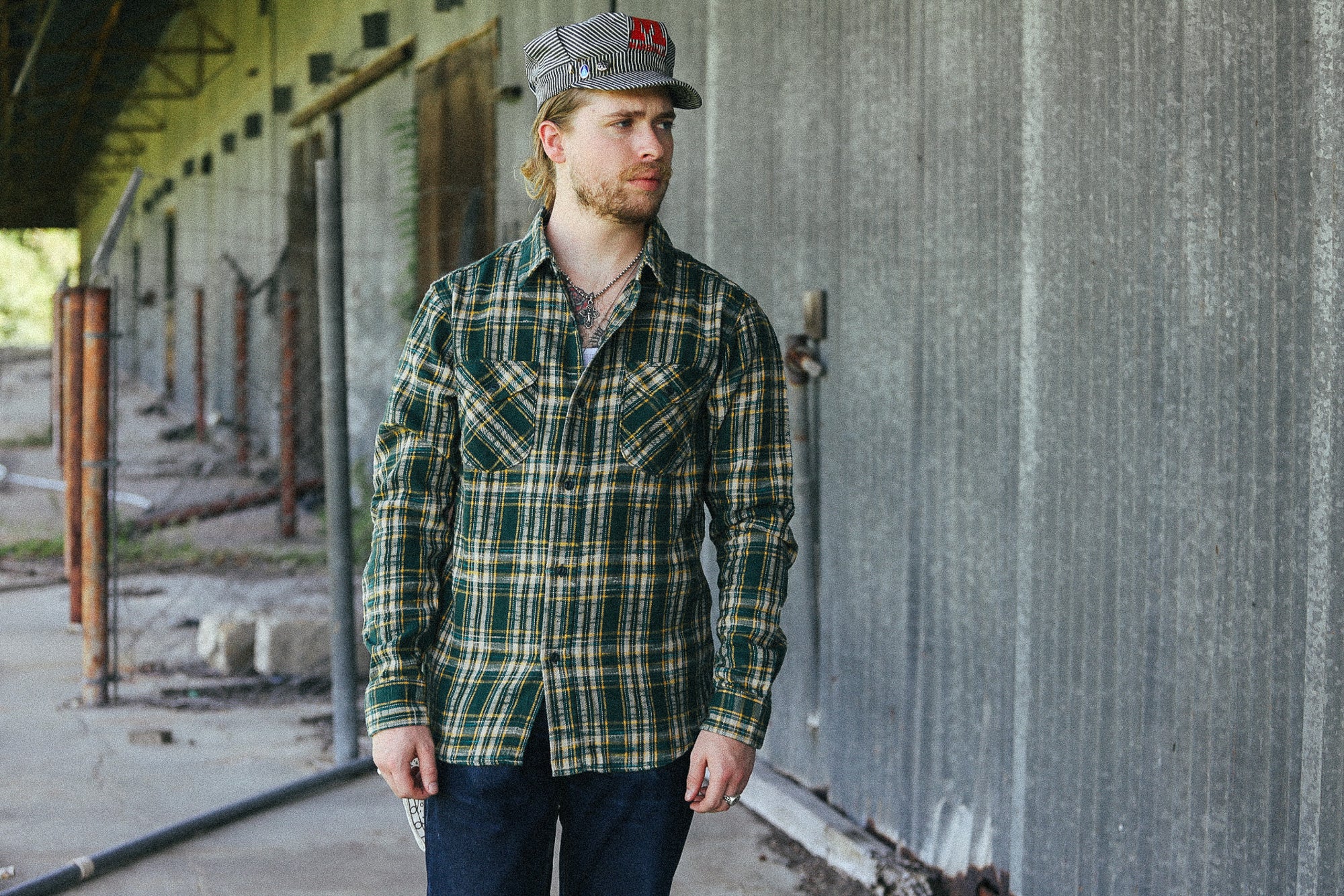 A man wearing a green and yellow plaid button-down flannel shirt, dark blue jeans, and a gray baseball cap with a red logo, standing in front of a corrugated metal wall.