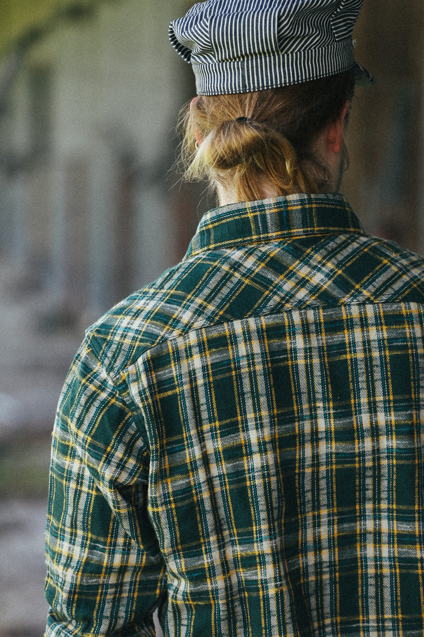 A person viewed from behind wearing a green, yellow, and white plaid flannel shirt and a striped black and white cap, standing in a softly blurred outdoor setting.